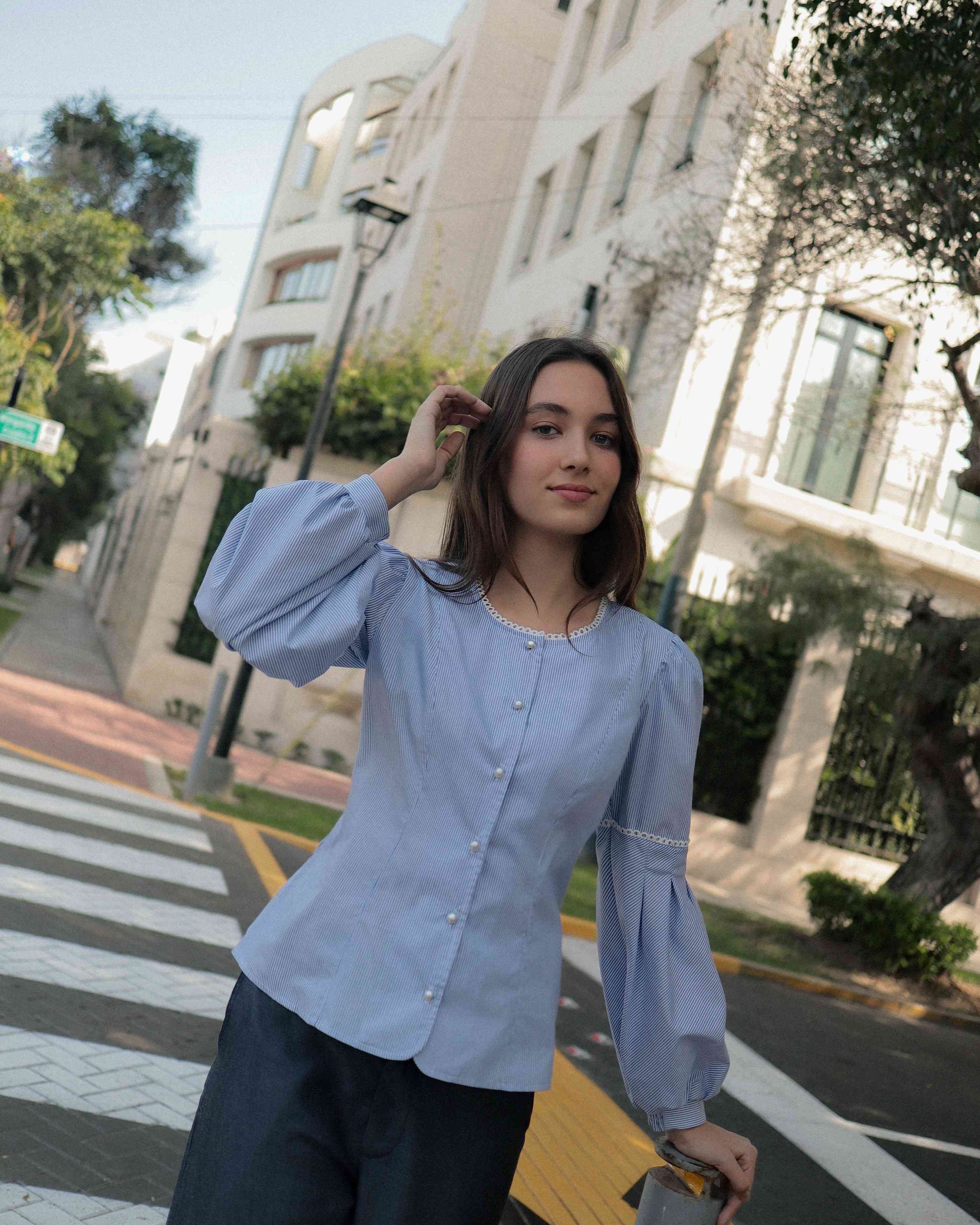 Mujer en la calle con blusa a rayas celeste ceñida al cuerpo con detalles tejidos, cuello redondo y mangas ligeramente voluminosas.