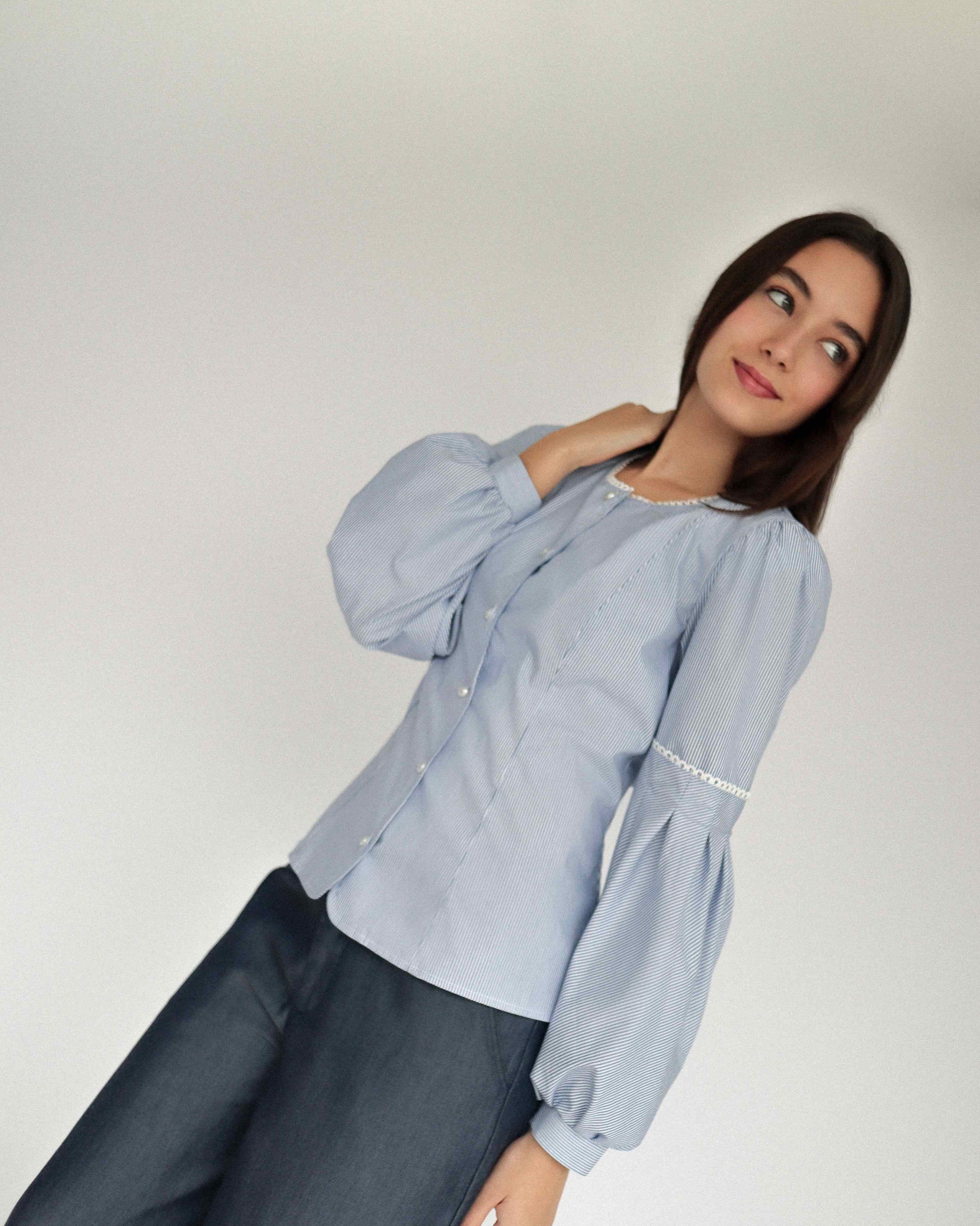 Mujer con blusa a rayas celeste con detalles tejidos, cuello redondo y mangas ligeramente voluminosas. Posando en fondo blanco.