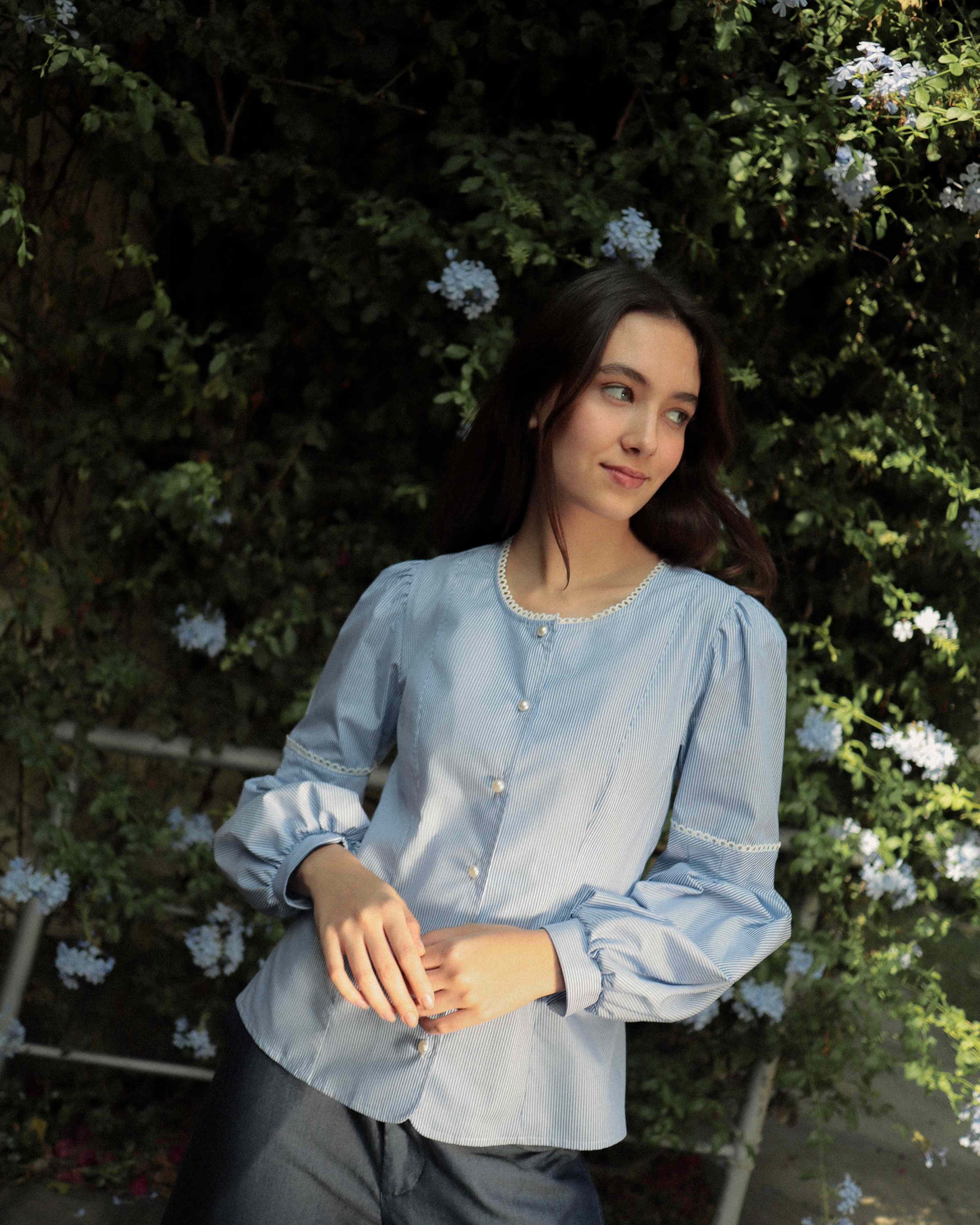 Mujer con blusa a rayas celeste con detalles tejidos, cuello redondo y mangas ligeramente voluminosas. Posando en fondo de flores.