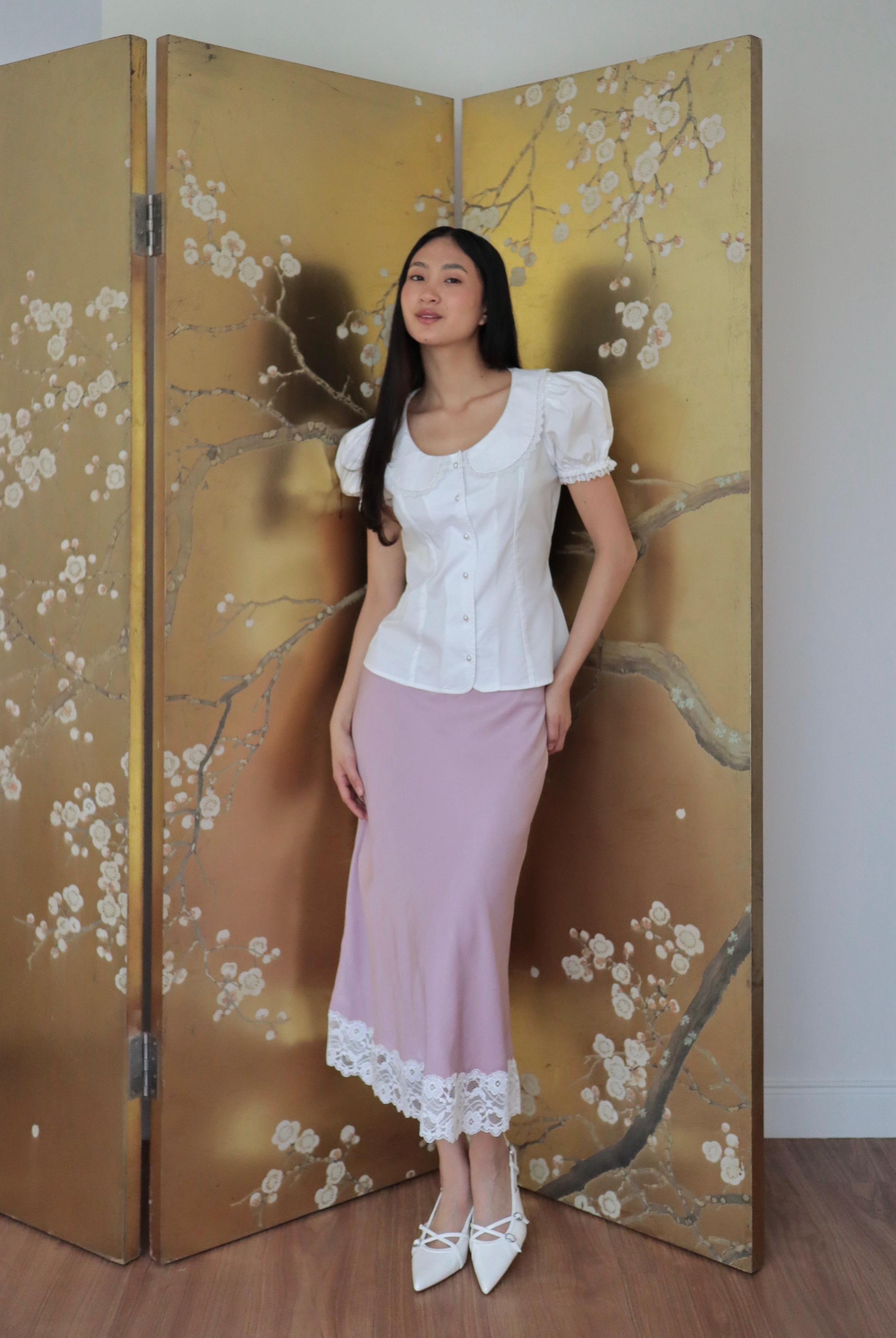 Mujer con blusa blanca de algodón romántica y falda rosa midi. Posando en fondo dorado con diseño floral