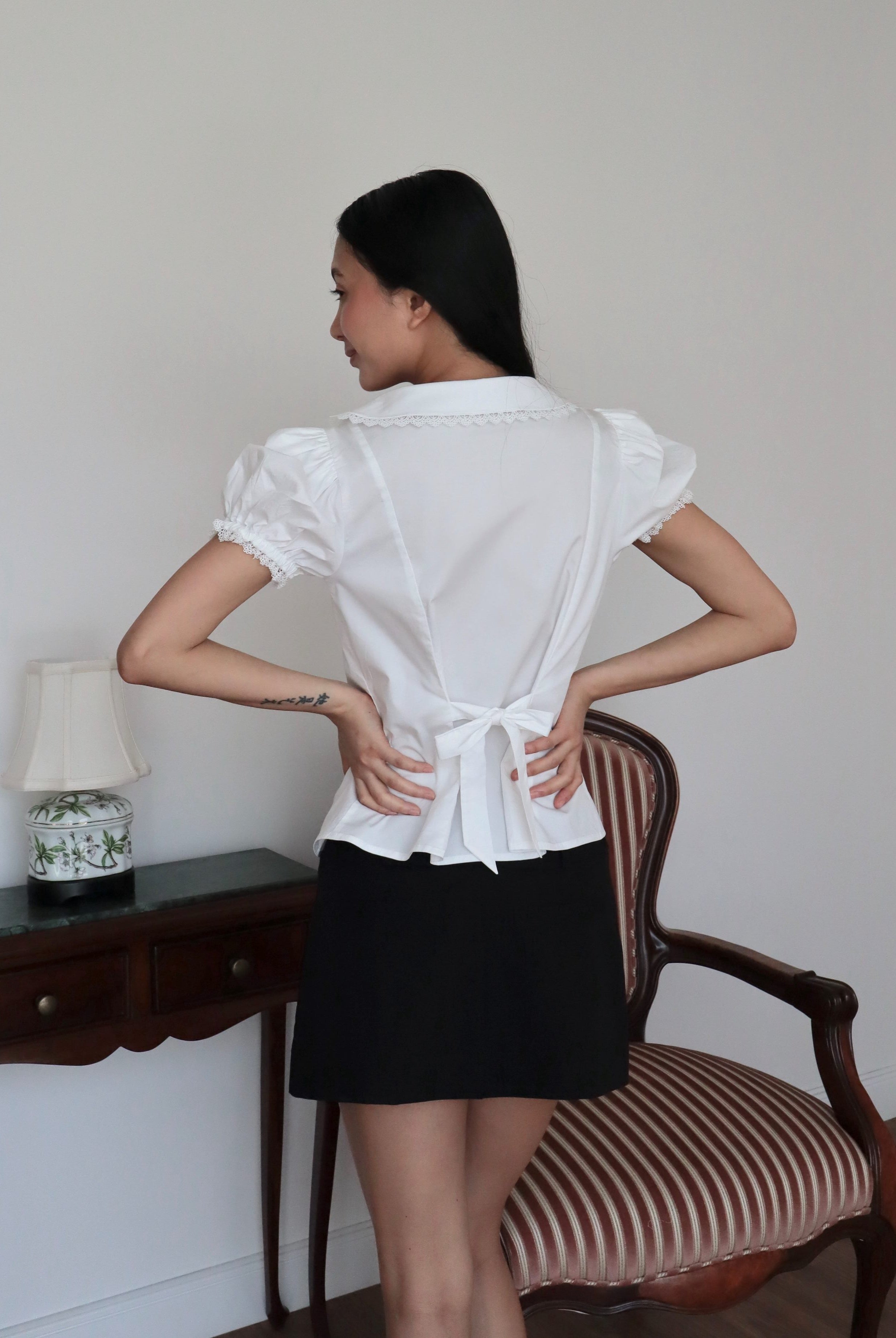 Mujer de espaldas con blusa blanca de algodón romántica con lazo y falda corta negra. Posando en fondo vintage