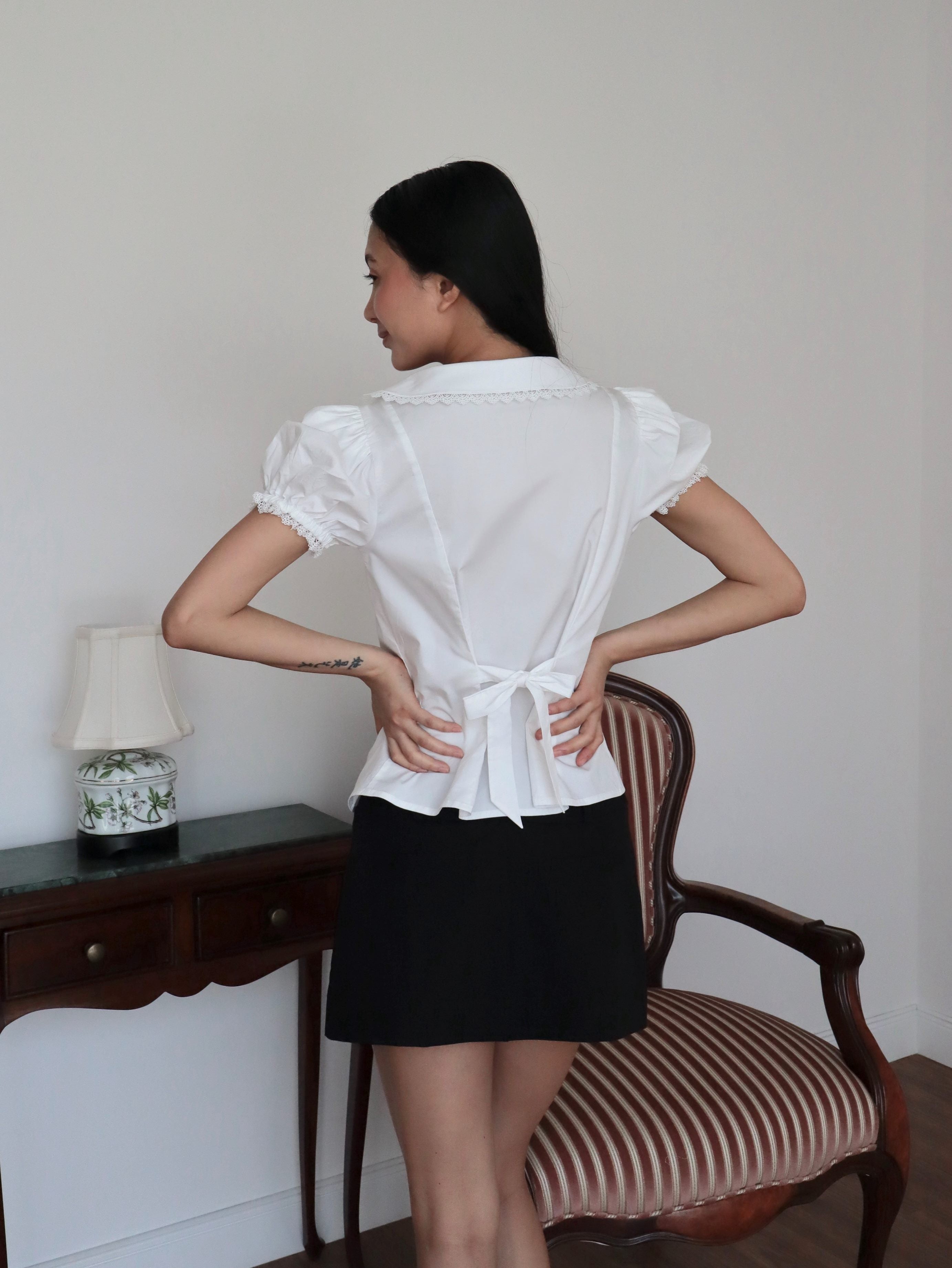 Mujer de espaldas con blusa blanca de algodón romántica con lazo y falda corta negra. Posando en fondo vintage
