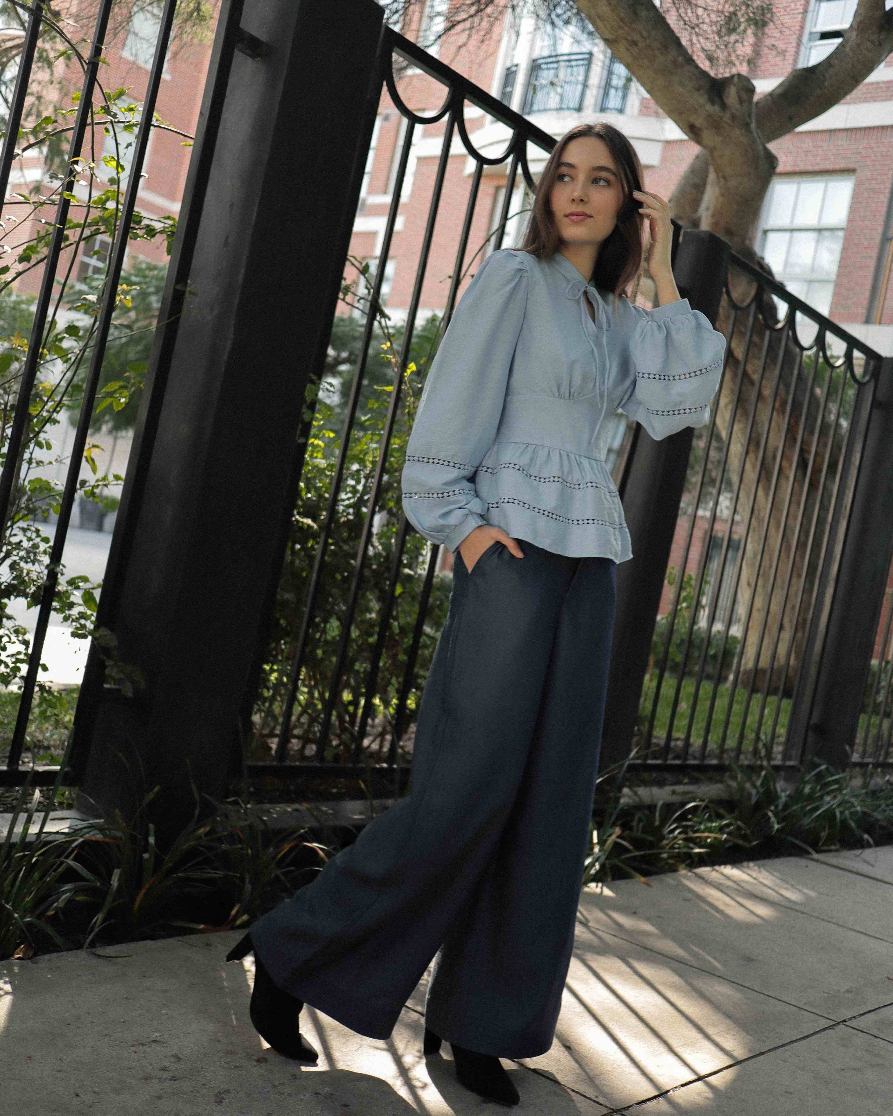 Mujer joven con blusa celeste con detalles tejidos, pantalones estilo denim y botas negras posando en fondo blanco