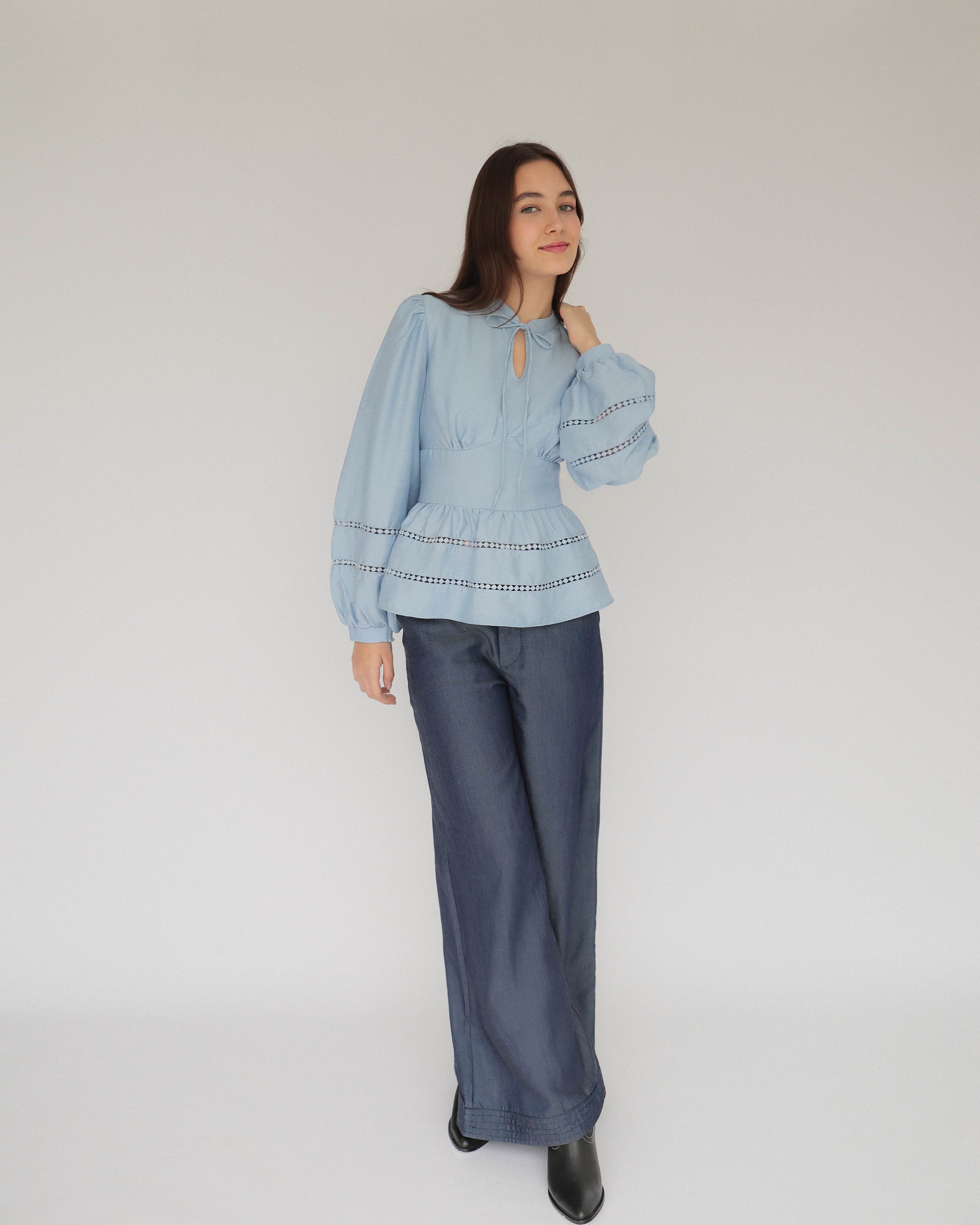 Mujer joven con blusa celeste con detalles tejidos, pantalones estilo denim y botas negras posando en fondo blanco