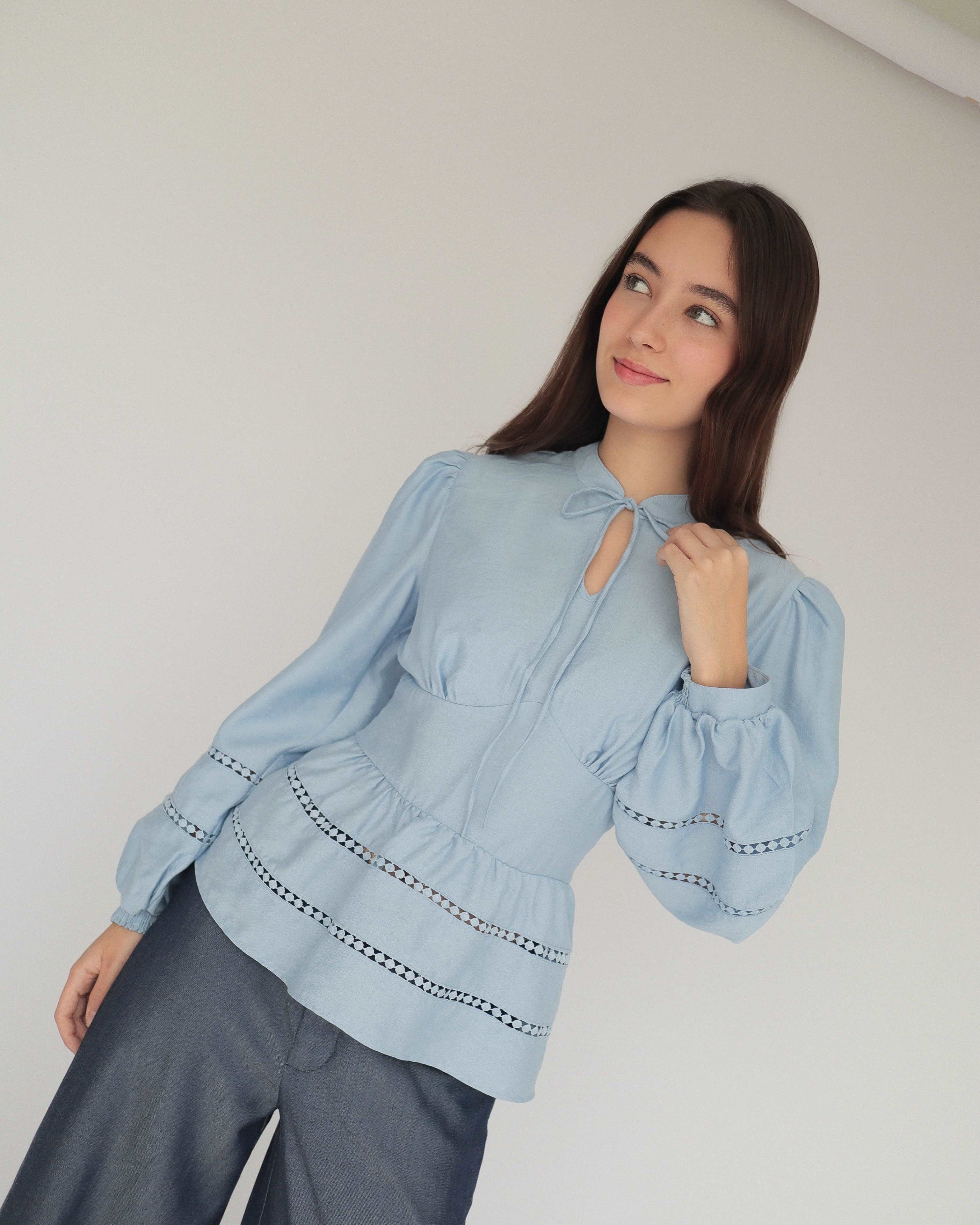 Mujer joven con blusa celeste con detalles tejidos y pantalones estilo denim posando en fondo blanco