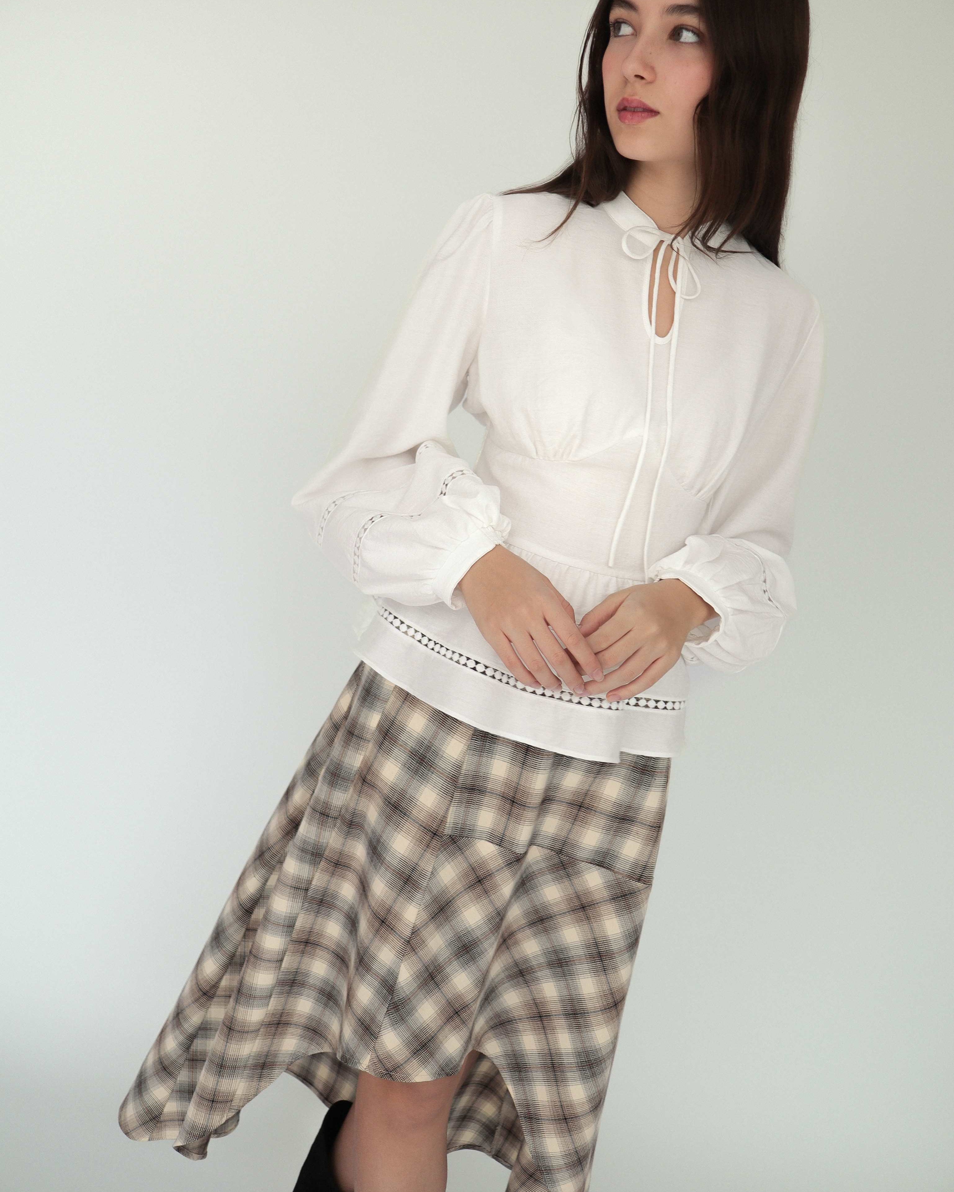 Mujer con blusa blanca sofisticada y elegante estilo peplum ceñida, con cuello alto y detalles tejidos. Lo combina con falda asimétrica a cuadros.