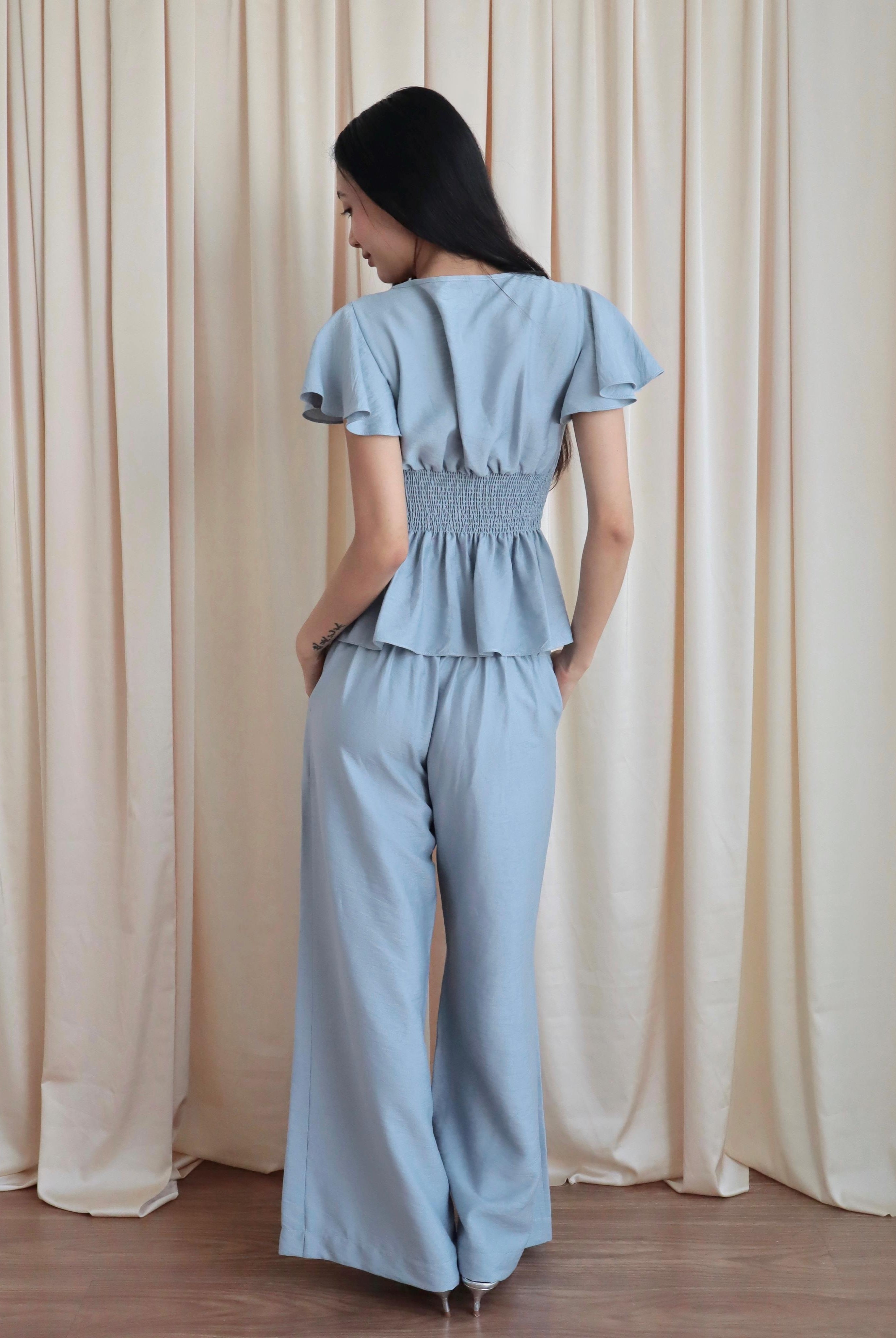 Mujer de espaldas usando blusa celeste romántica con bobos en mangas, lazo y corte que marca la cintura. Usa un pantalón recto celeste. Posando en fondo amarillo pastel.