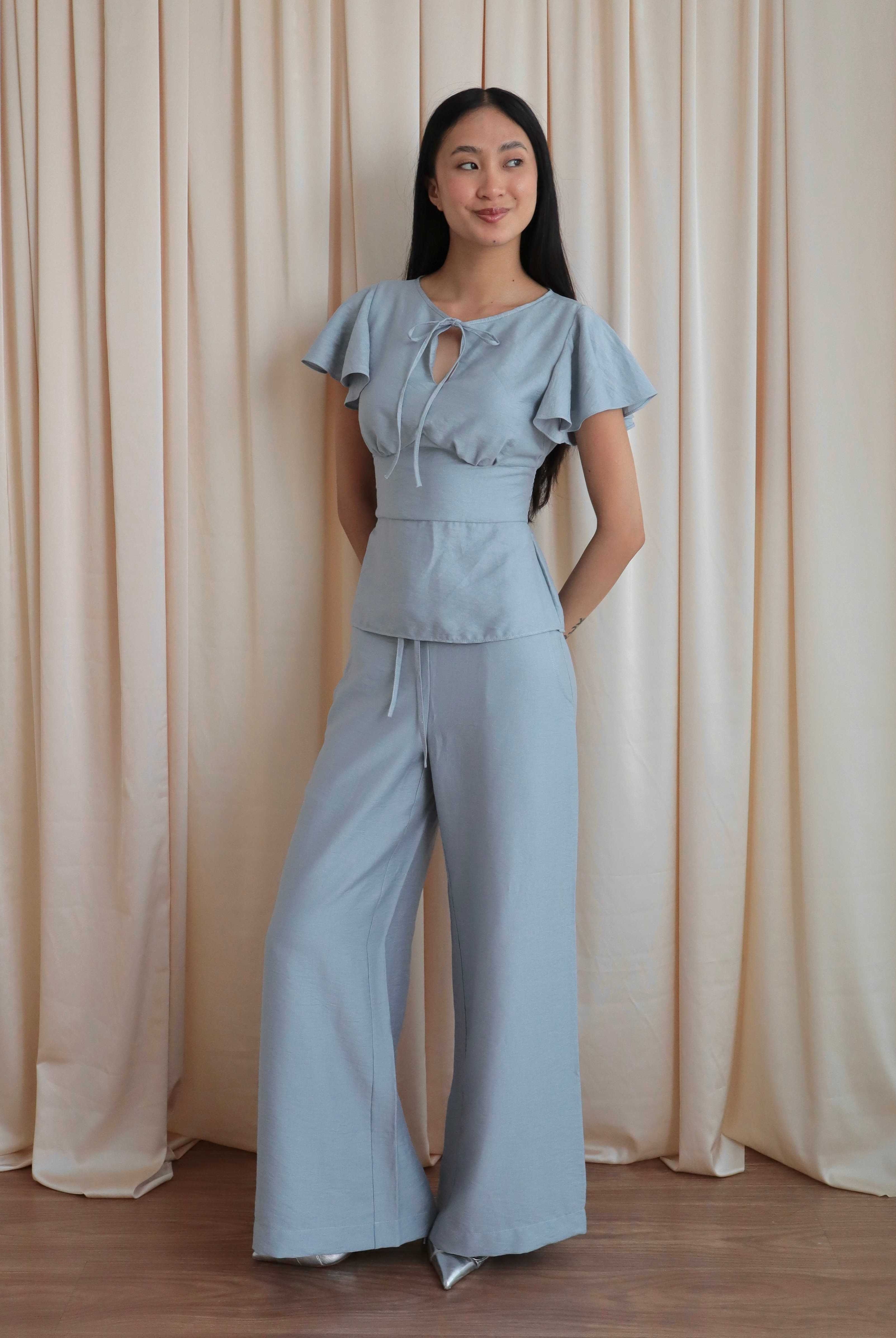 Mujer con blusa celeste romántica con bobos en mangas y lazo. Pantalón recto celeste. Posando en fondo amarillo pastel.