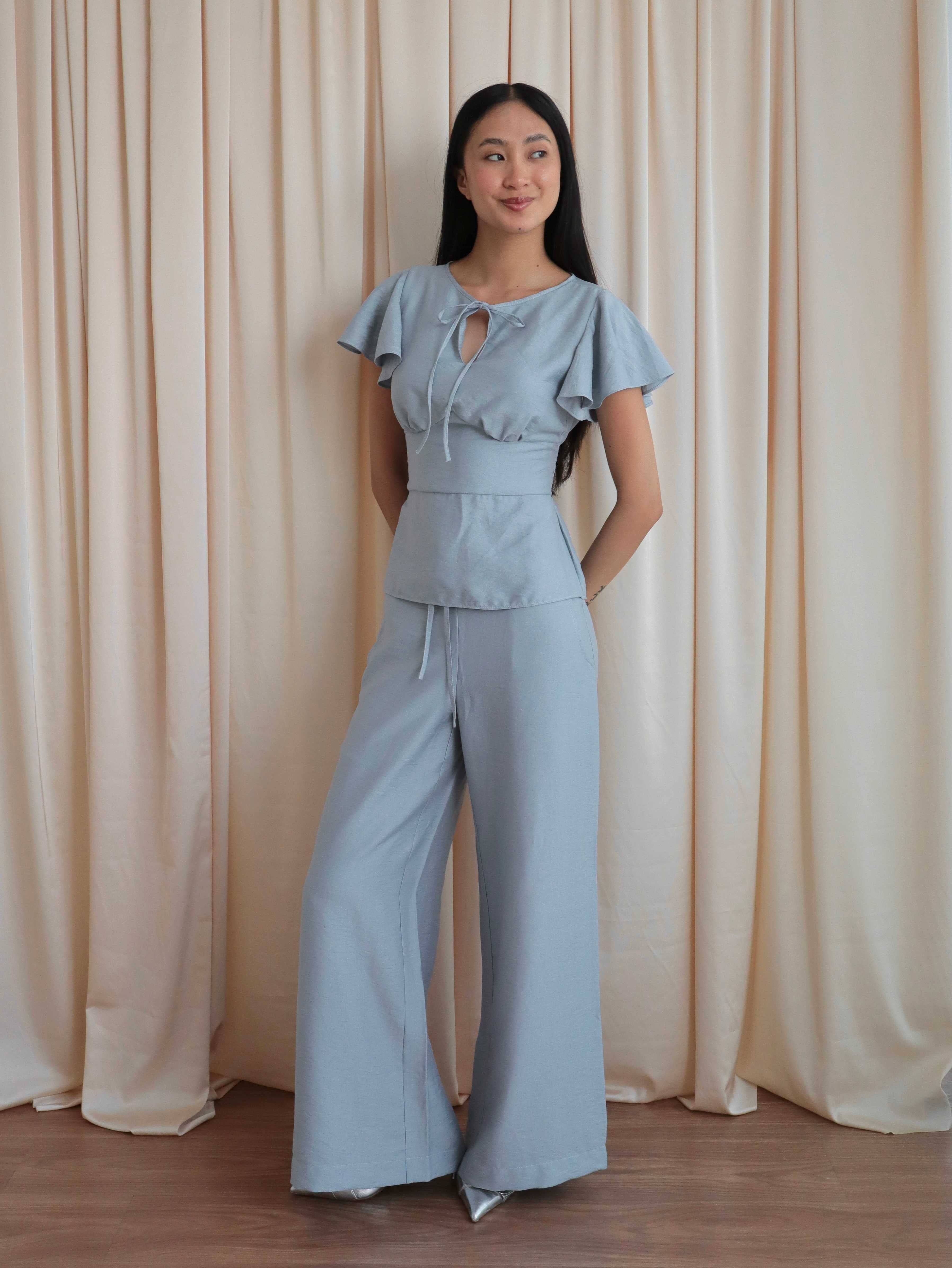 Mujer con blusa celeste romántica con bobos en mangas y lazo. Pantalón recto celeste. Posando en fondo amarillo pastel.