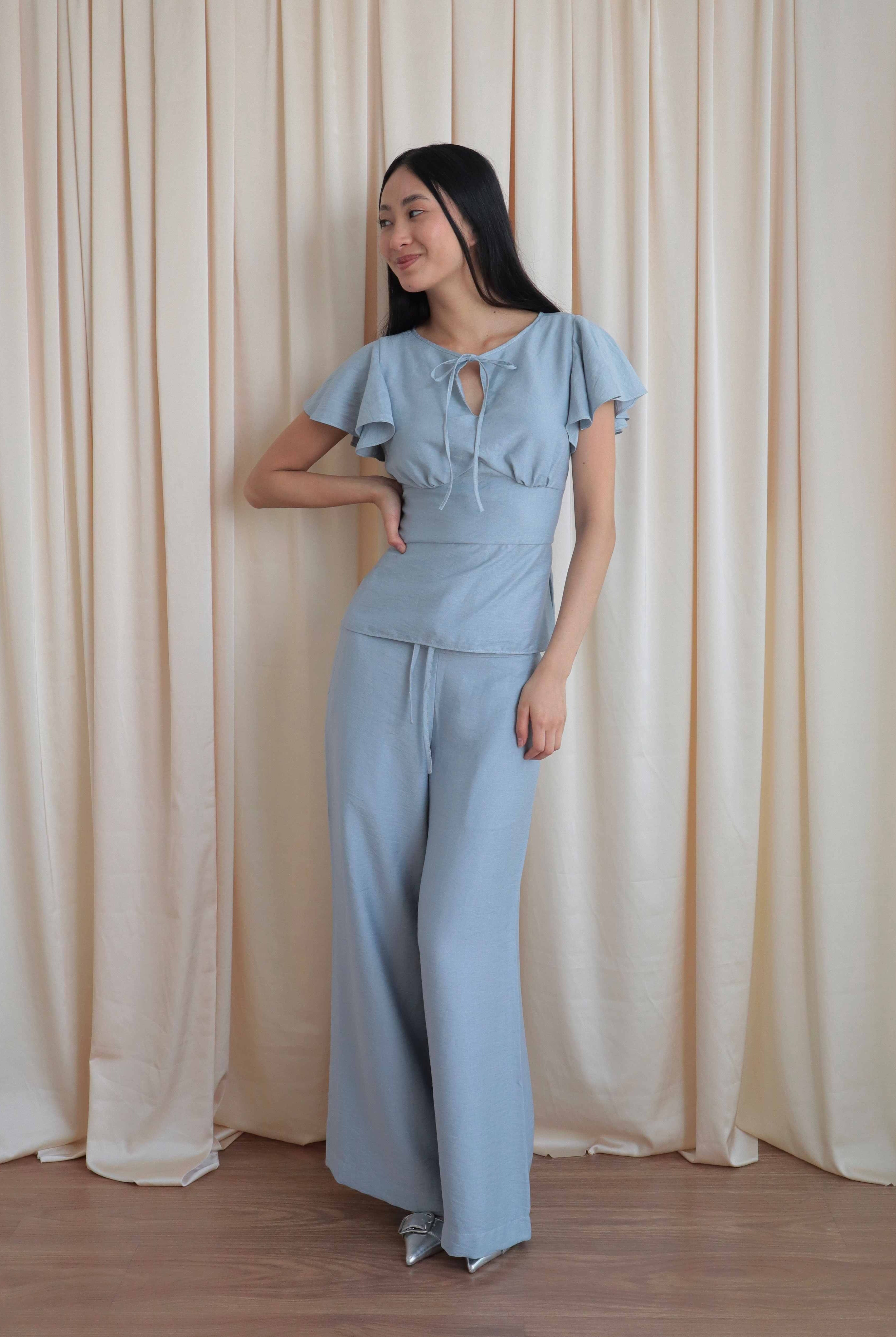 Mujer con blusa celeste romántica con bobos en mangas y lazo. Pantalón recto celeste. Posando en fondo amarillo pastel.