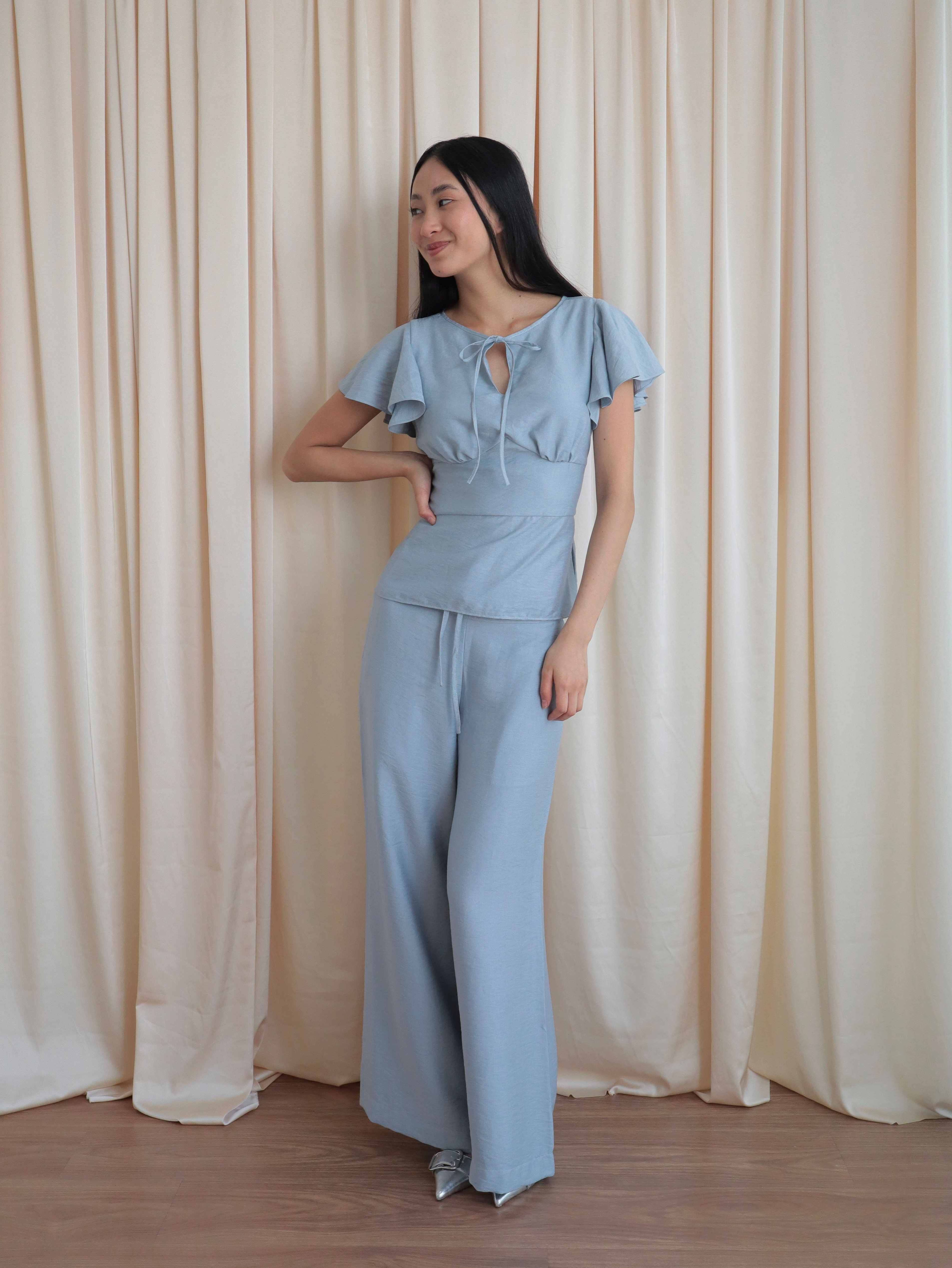 Mujer con blusa celeste romántica con bobos en mangas y lazo. Pantalón recto celeste. Posando en fondo amarillo pastel.