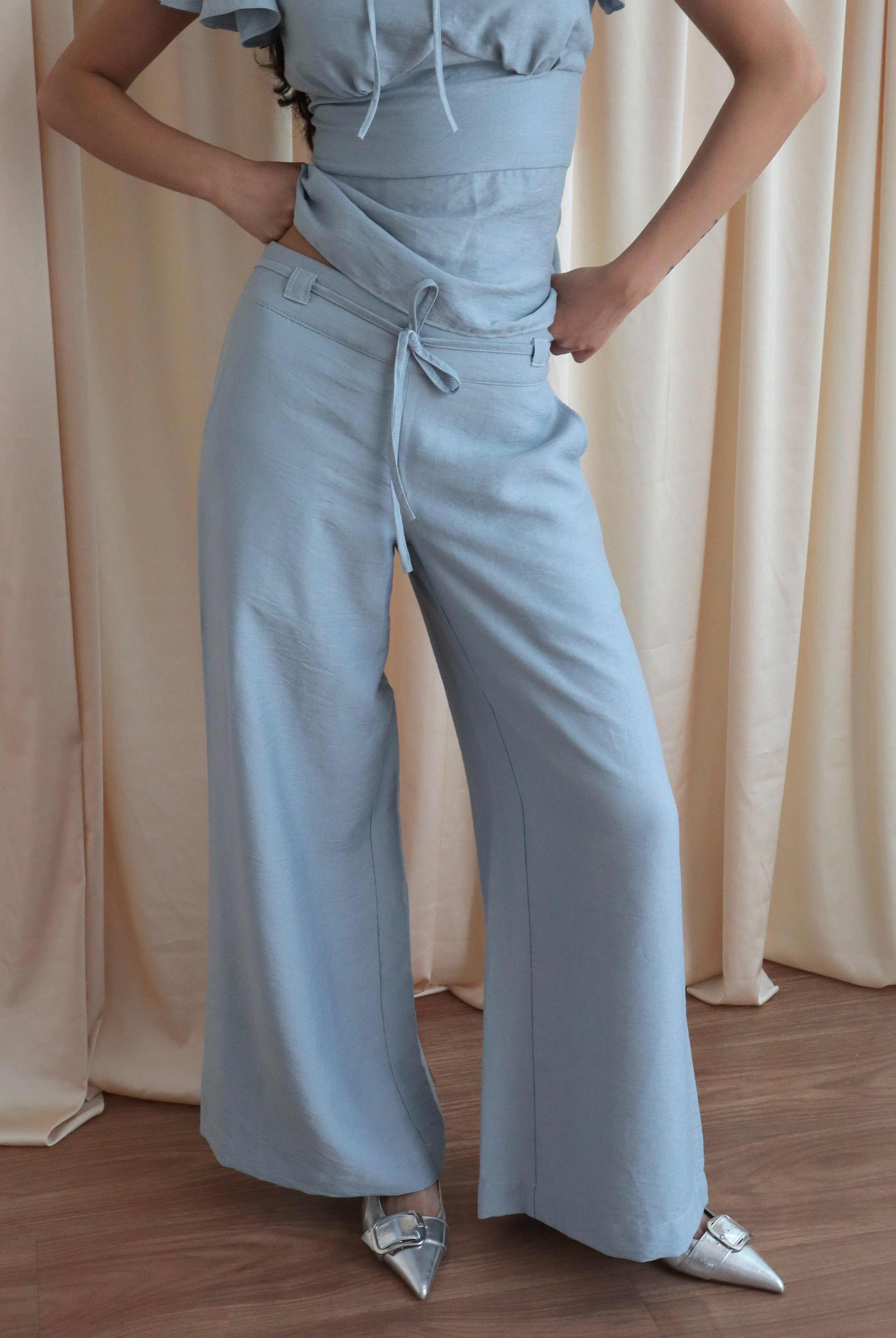 Mujer usando pantalón recto a la cadera celeste con lazo en pretina. Posando en fondo amarillo pastel.