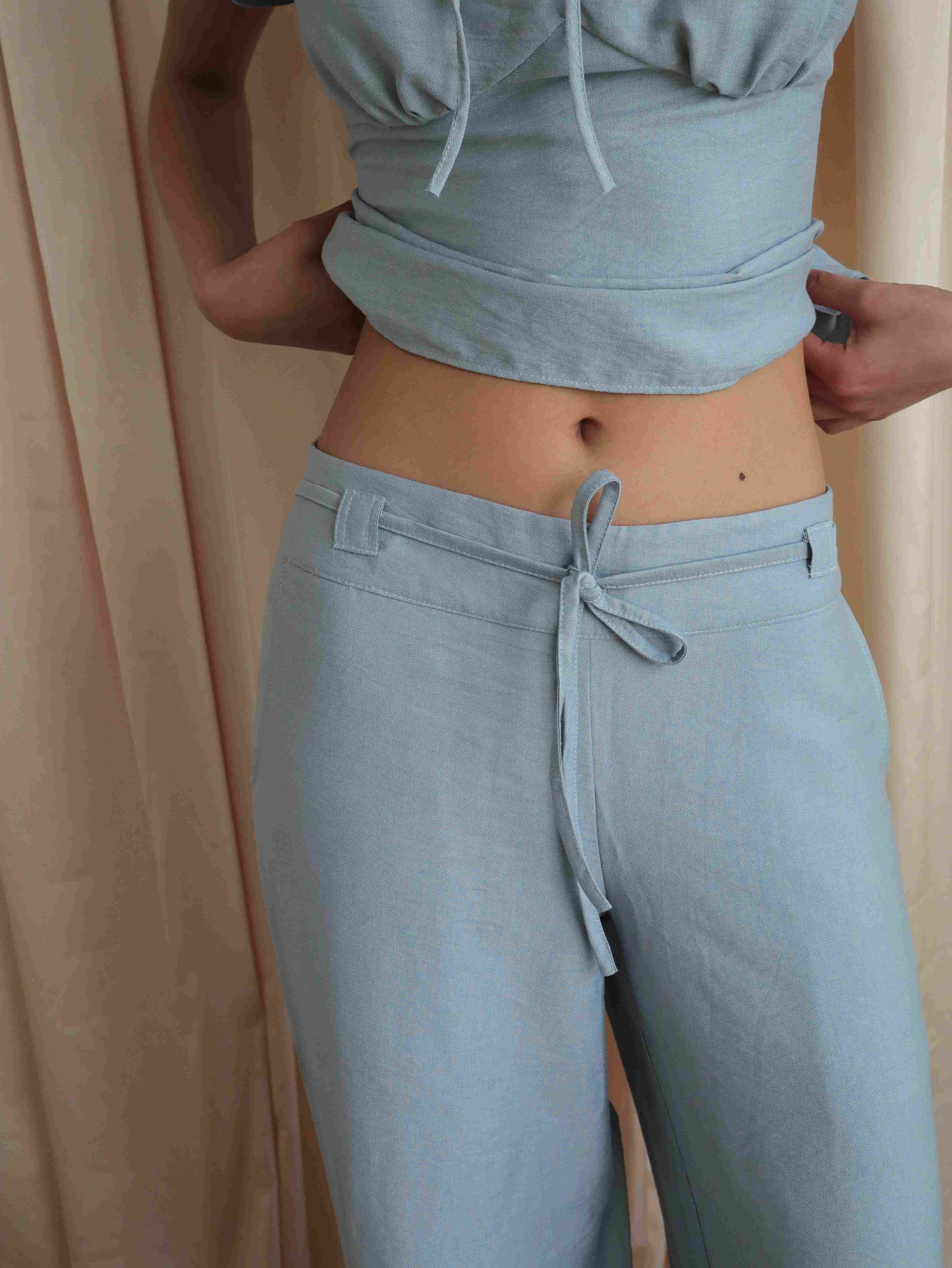 Mujer usando pantalón recto a la cadera celeste con lazo en pretina. Posando en fondo amarillo pastel.