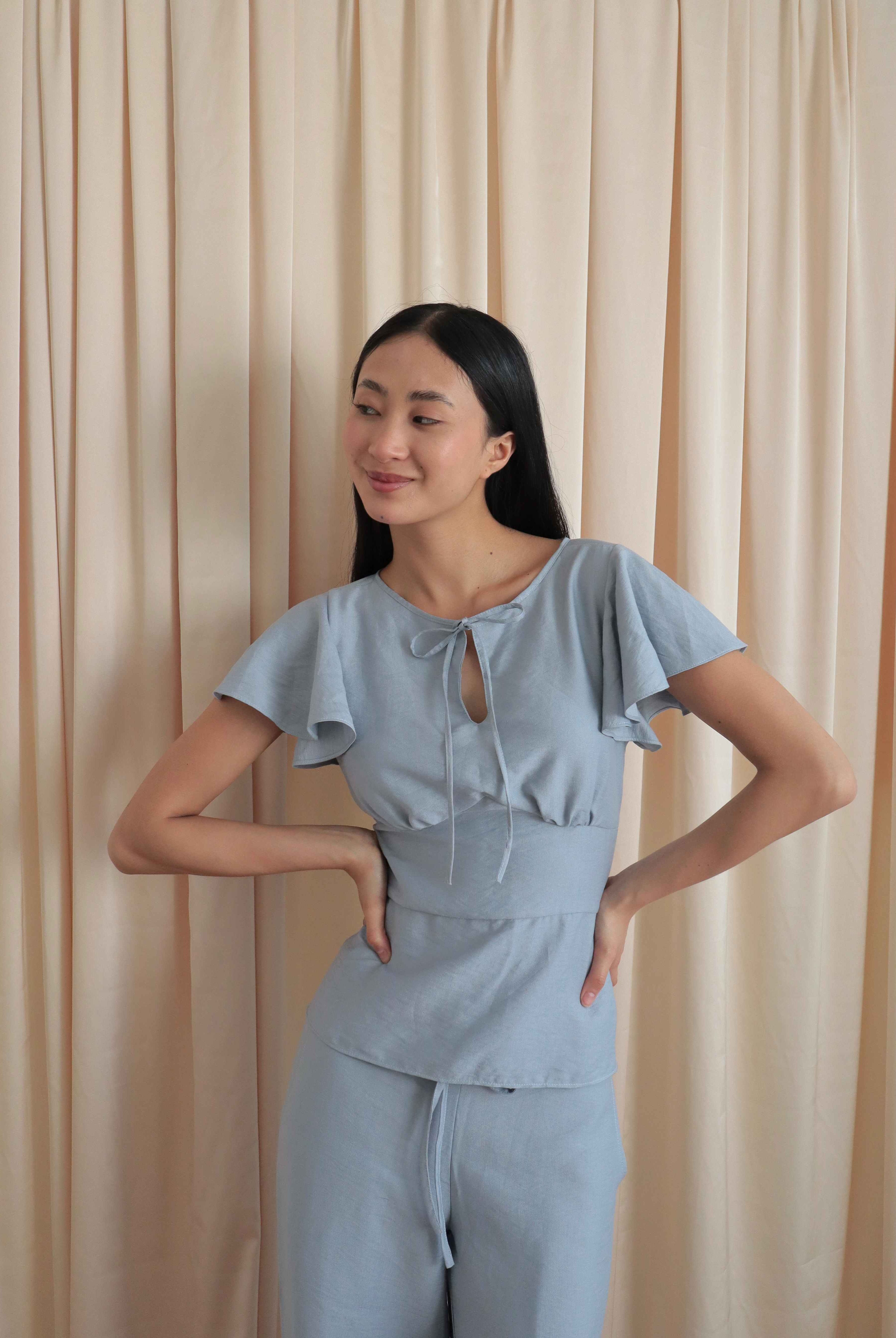 Mujer con blusa celeste romántica con bobos en mangas, lazo y corte que marca la cintura. Posando en fondo amarillo pastel.