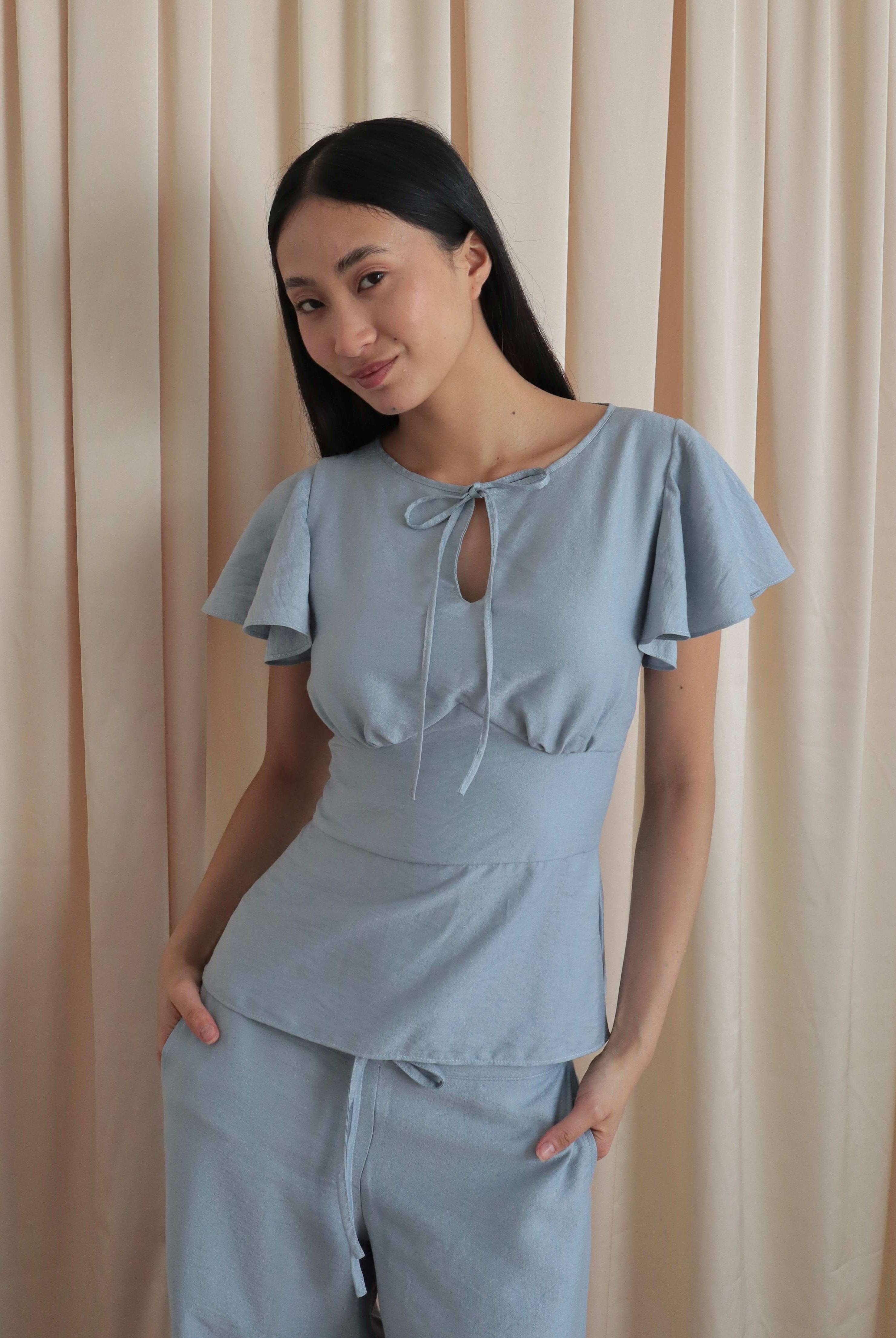 Mujer con blusa celeste romántica con bobos en mangas, lazo y corte que marca la cintura. Posando en fondo amarillo pastel.