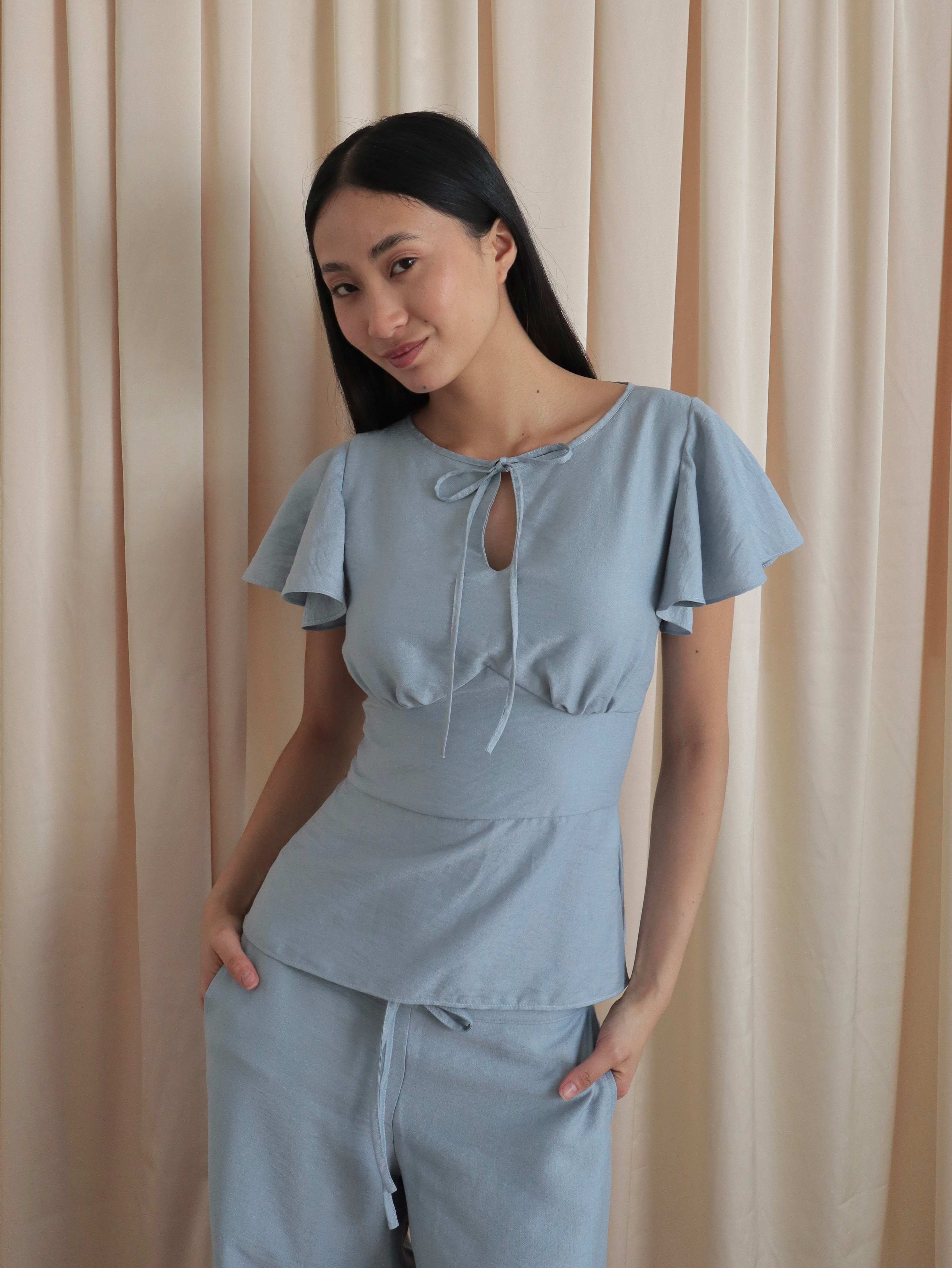 Mujer con blusa celeste romántica con bobos en mangas, lazo y corte que marca la cintura. Posando en fondo amarillo pastel.