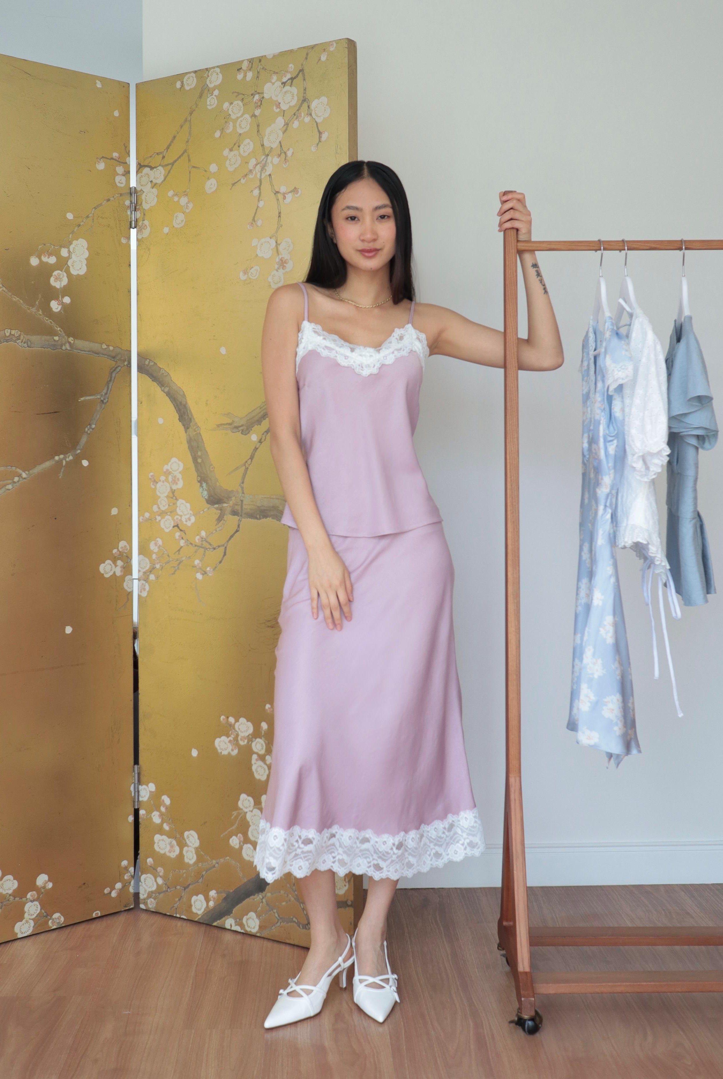 Mujer usando conjunto de top y falda midi lencero rosa romántico en fondo floral y rack de ropa