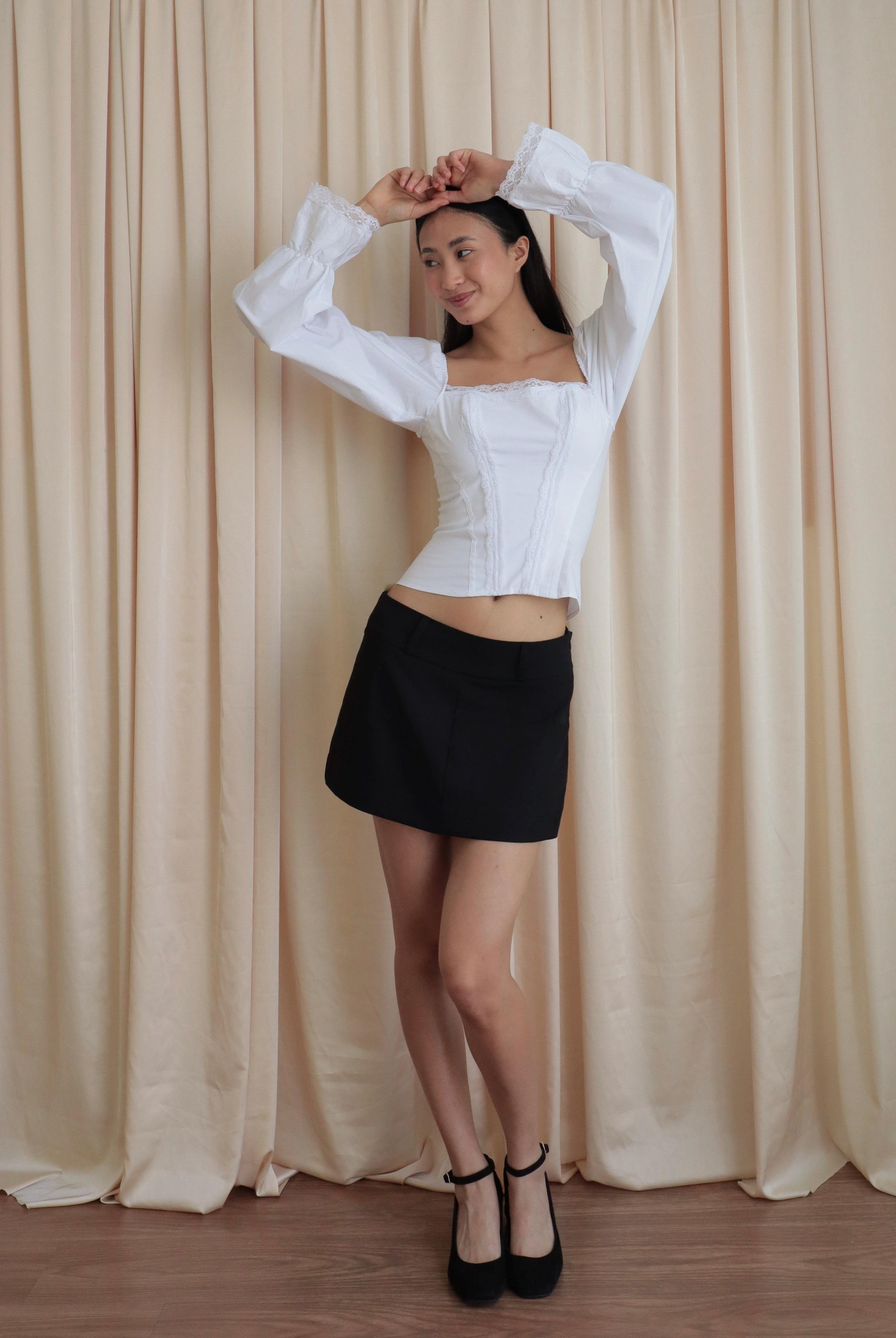 Mujer usando corset de algodón, mangas largas y blondas, usando falda negra corta con ballerinas. Posando en fondo amarillo pastel.