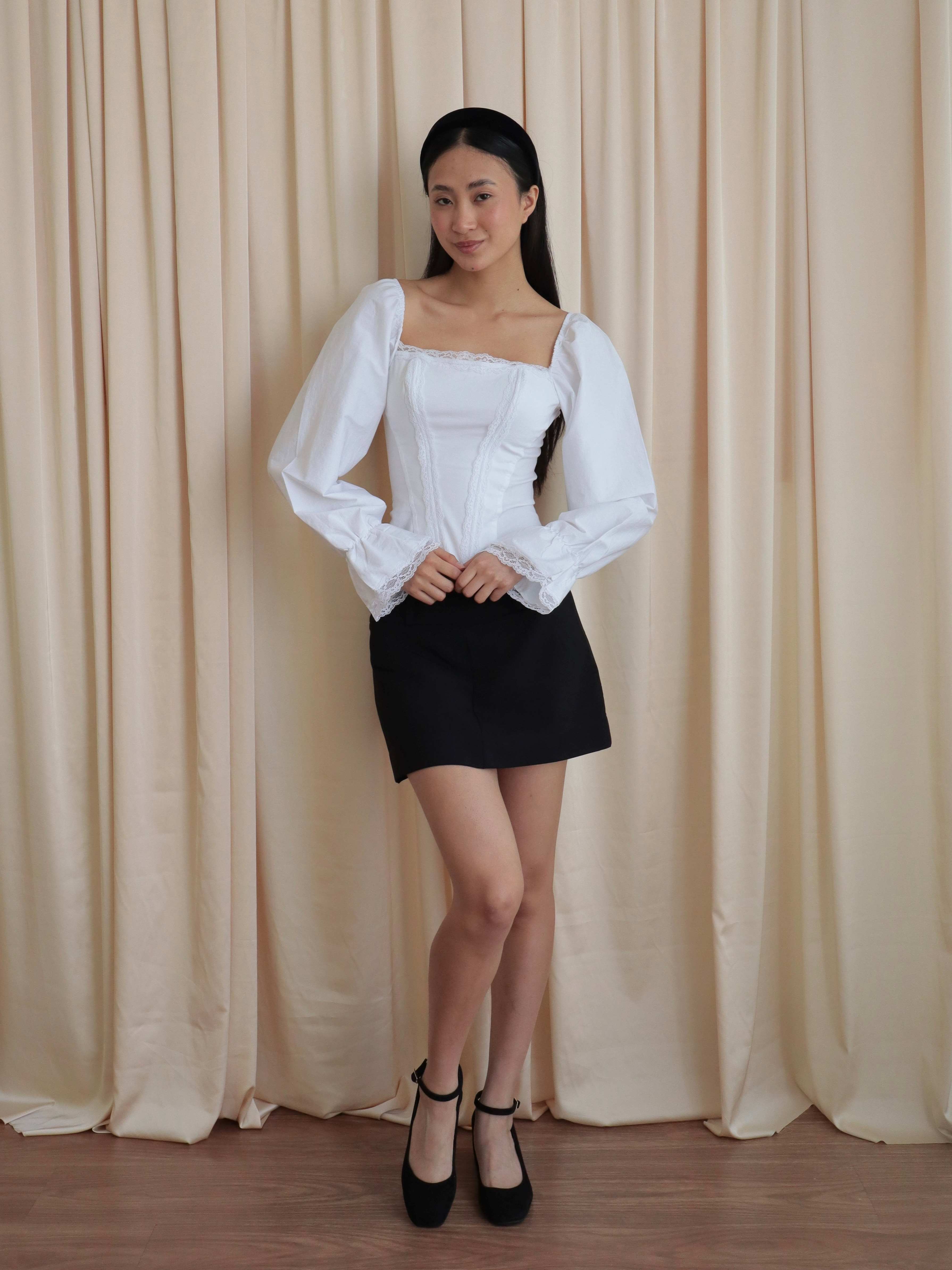 Mujer usando corset de algodón, mangas largas y blondas, usando falda negra corta con ballerinas. Posando en amarillo pastel.