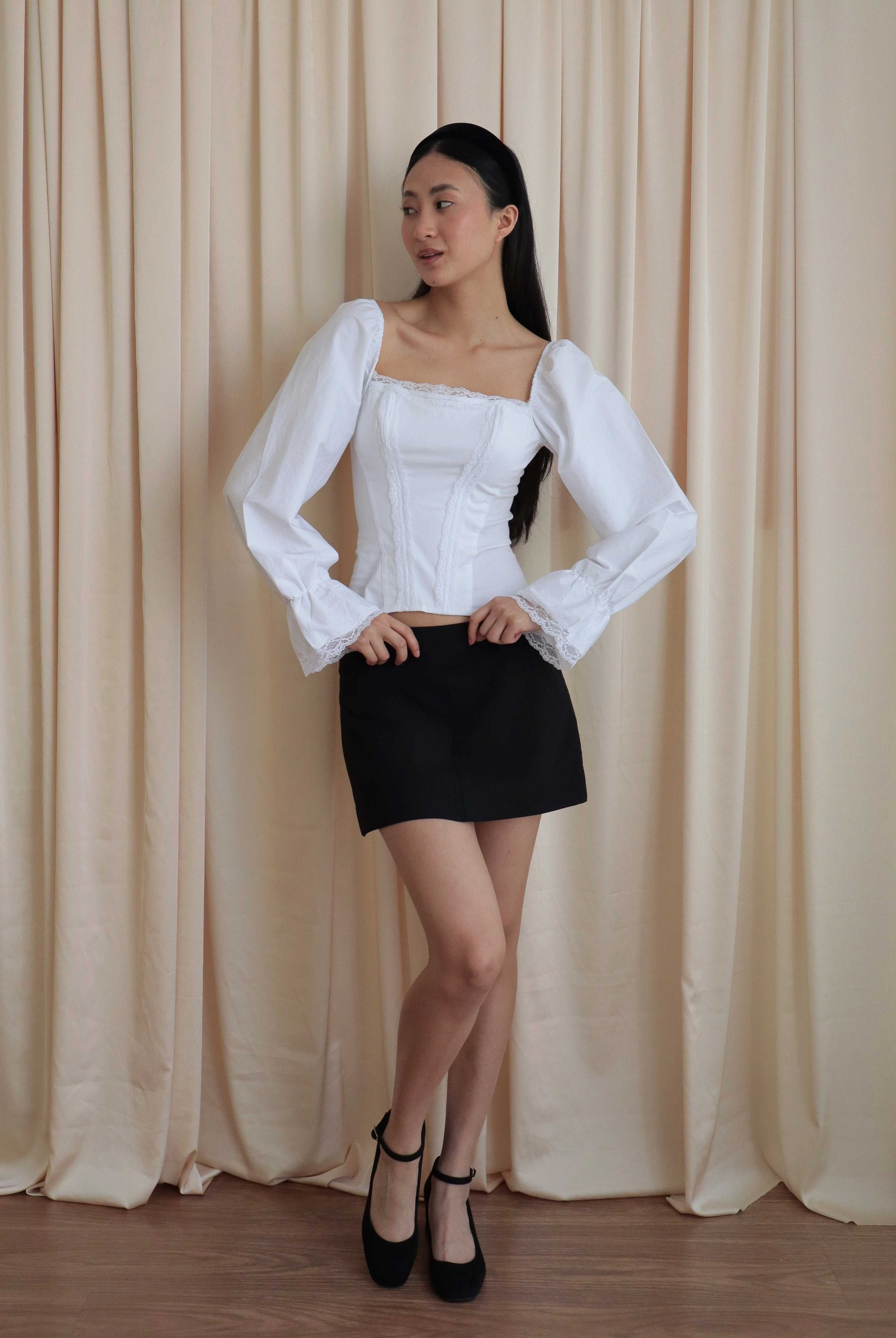 Mujer usando corset de algodón, mangas largas y blondas, usando falda negra corta con ballerinas. Posando en fondo amarillo pastel.