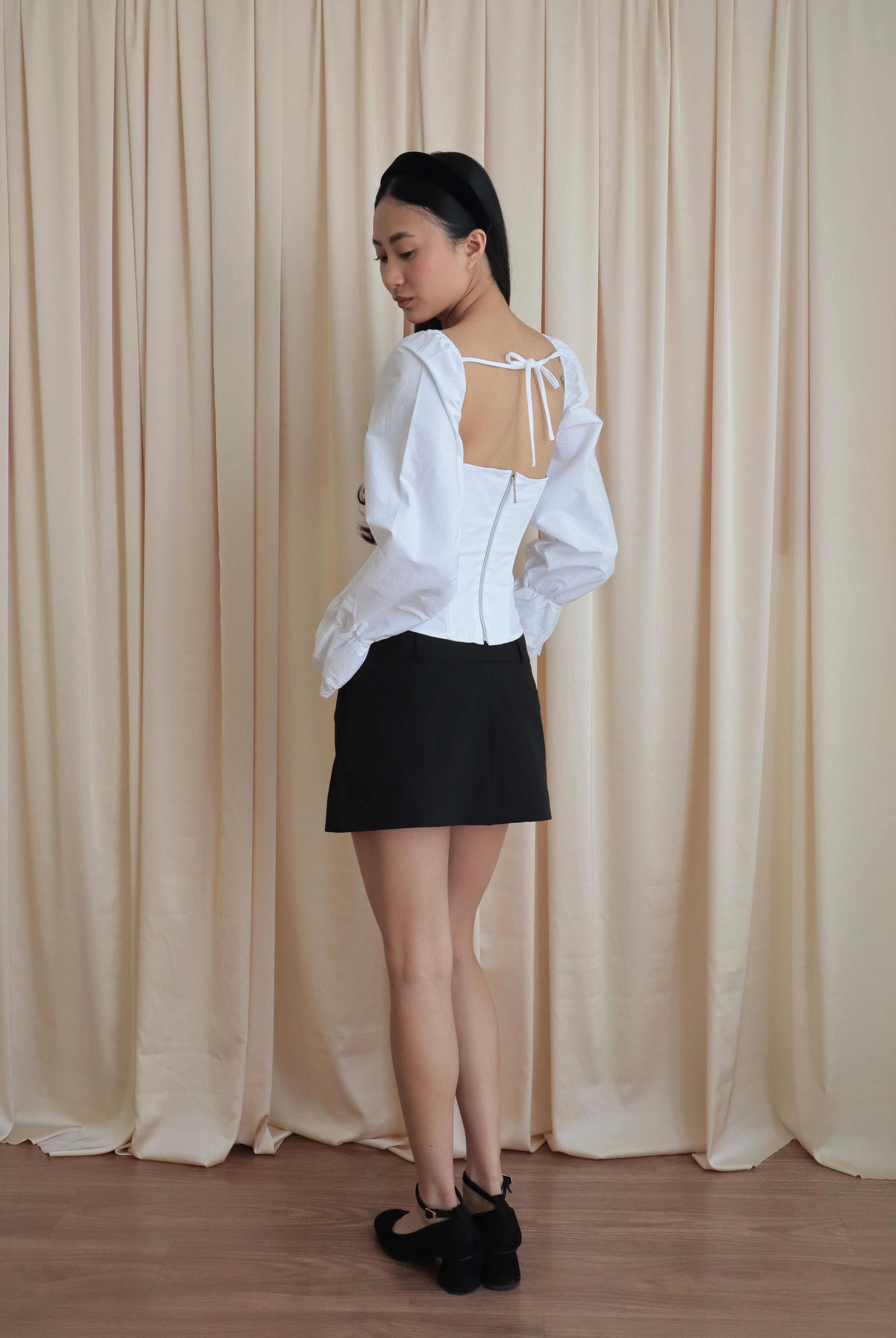 Mujer usando corset de algodón, mangas largas y blondas, usando falda negra corta con ballerinas. Posando en fondo amarillo pastel.