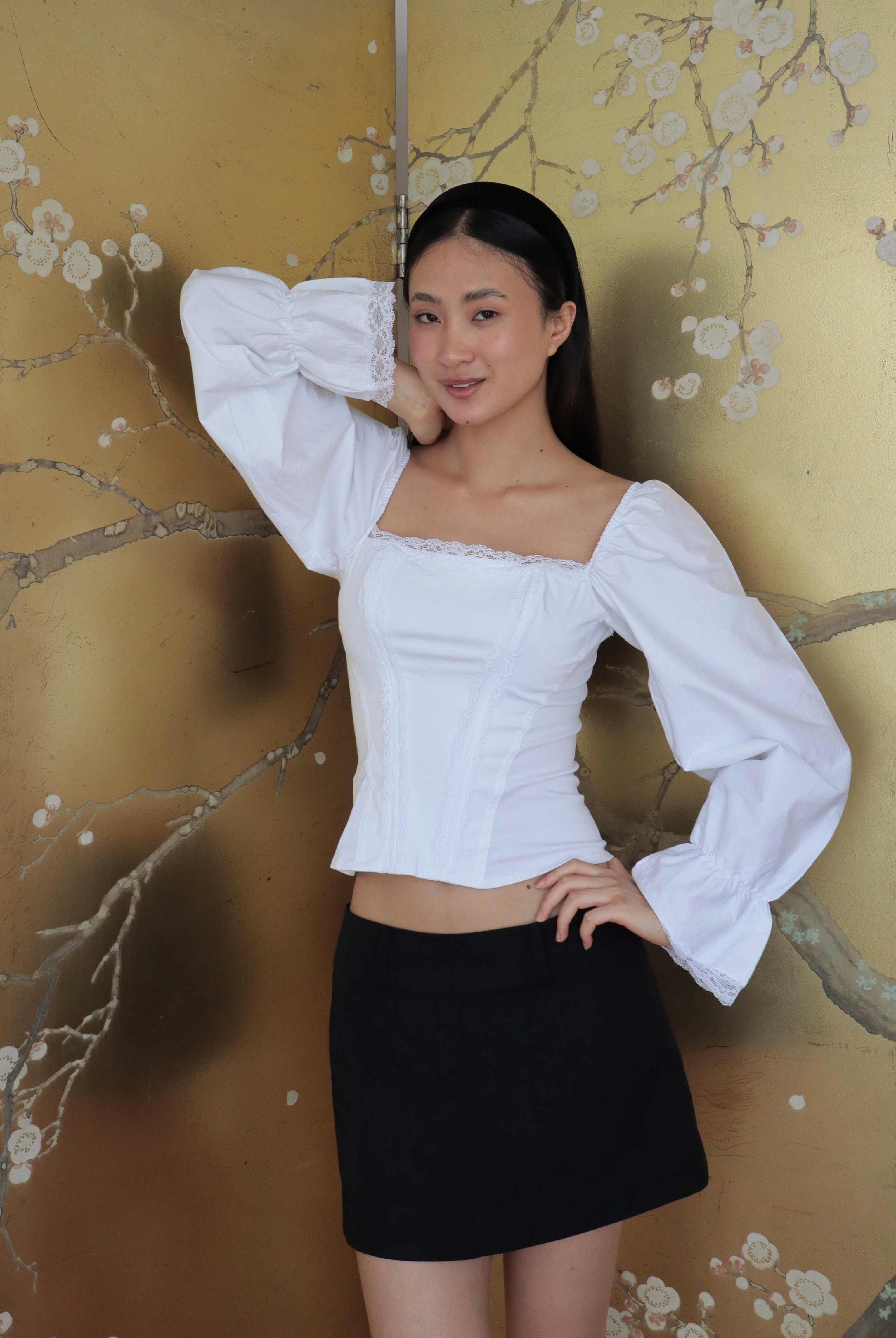 Mujer usando corset de algodón, mangas largas y blondas, usando falda negra corta. Posando en fondo floral.