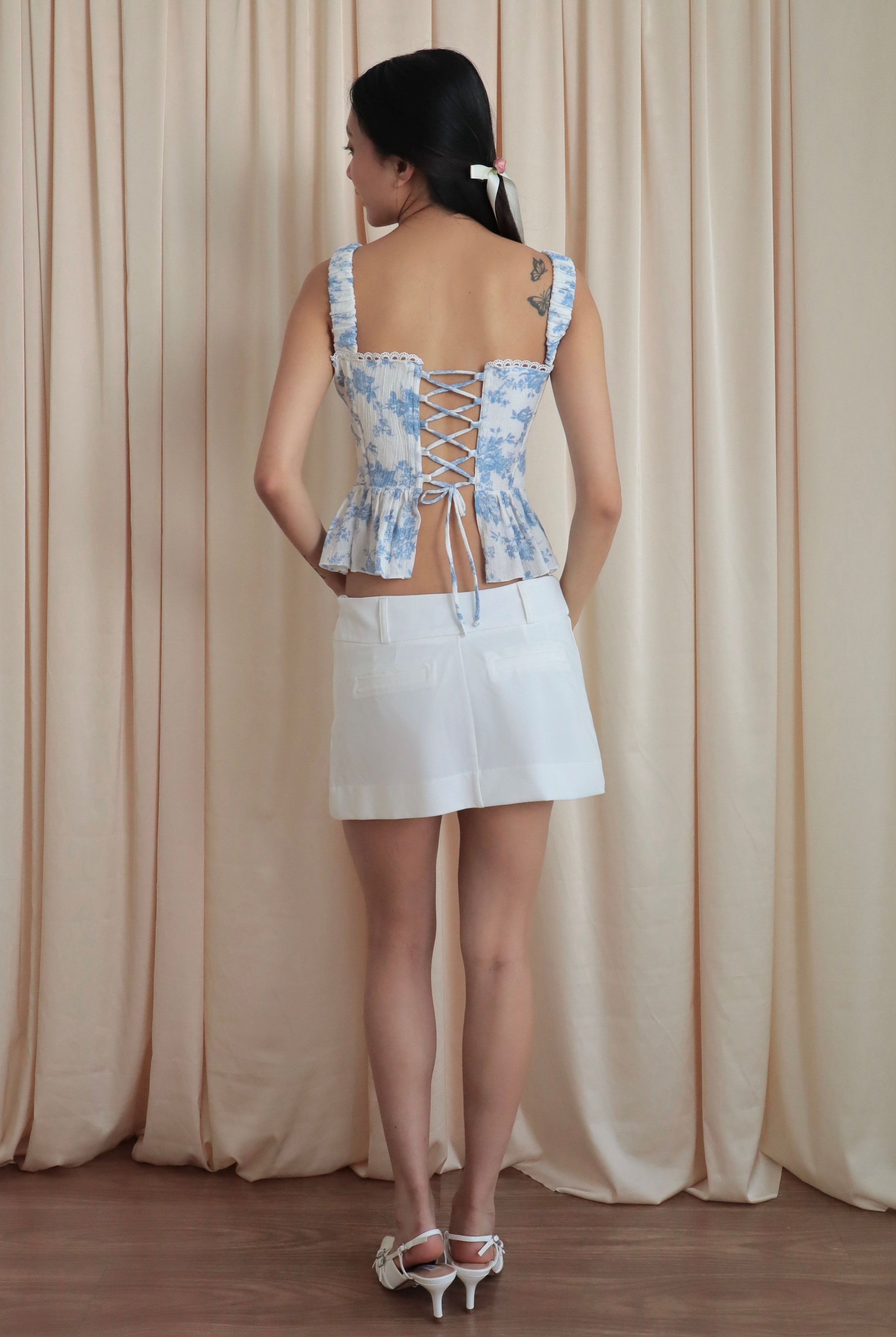 Mujer de espalda con corset romántico estampado flores celestes con pasadores y falda blanca. Posando en fondo amarillo pastel.