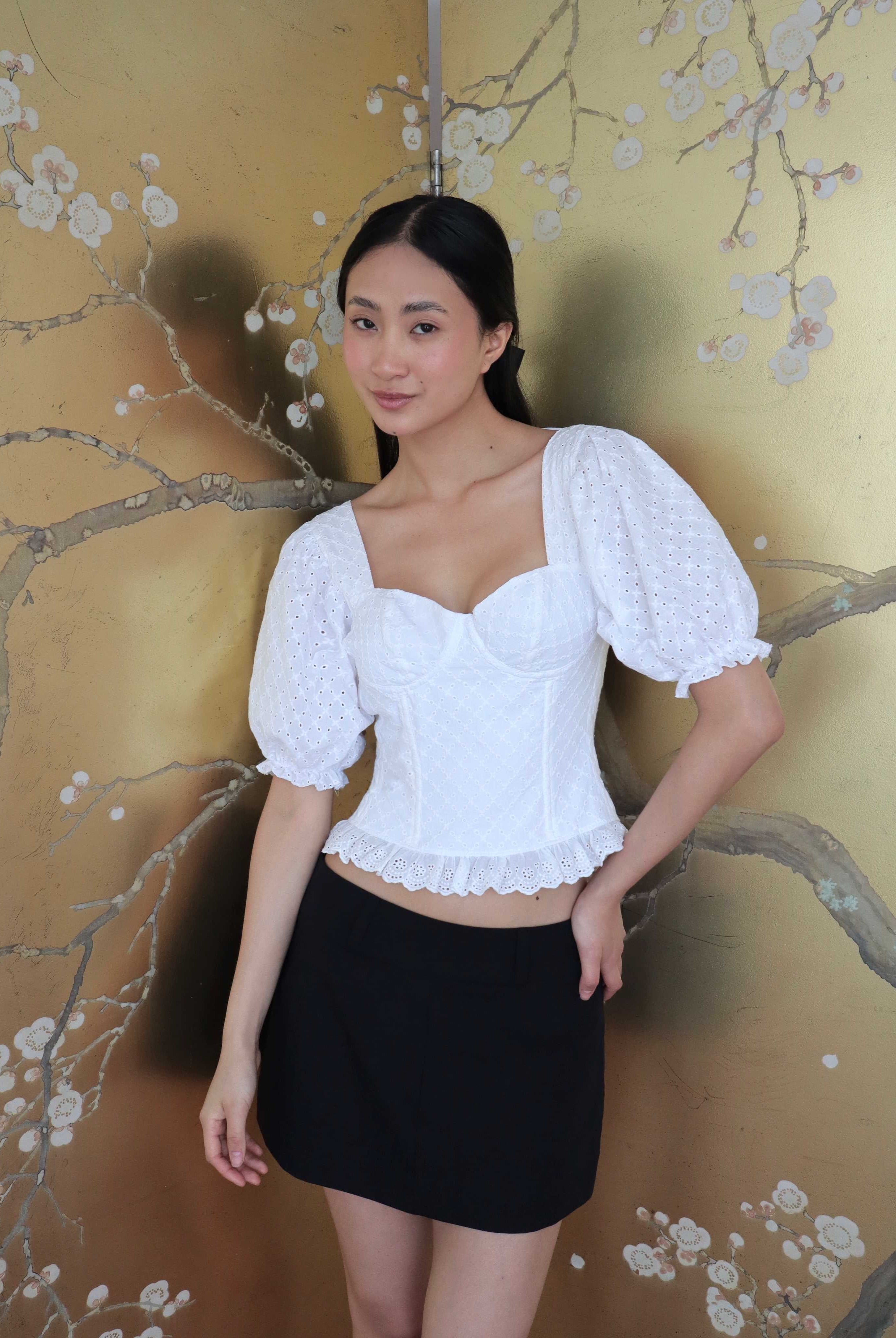 Mujer con corset broderie con escote corazón, mangas abullonadas y blonda usando falda negra corta. Posando en fondo floral.