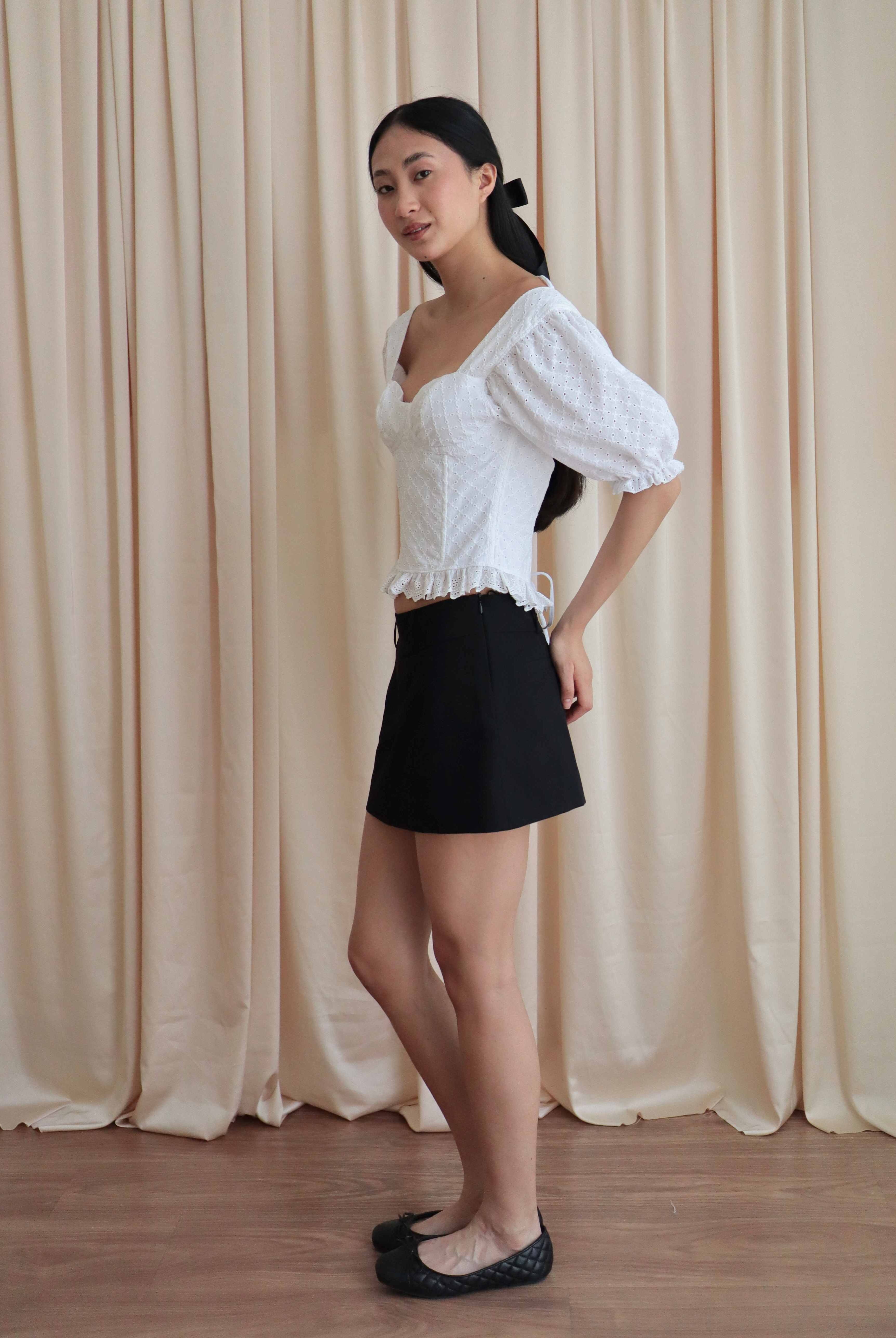 Mujer con corset broderie con escote corazón, mangas abullonadas y blonda usando falda negra corta con ballerinas. Posando en amarillo pastel.