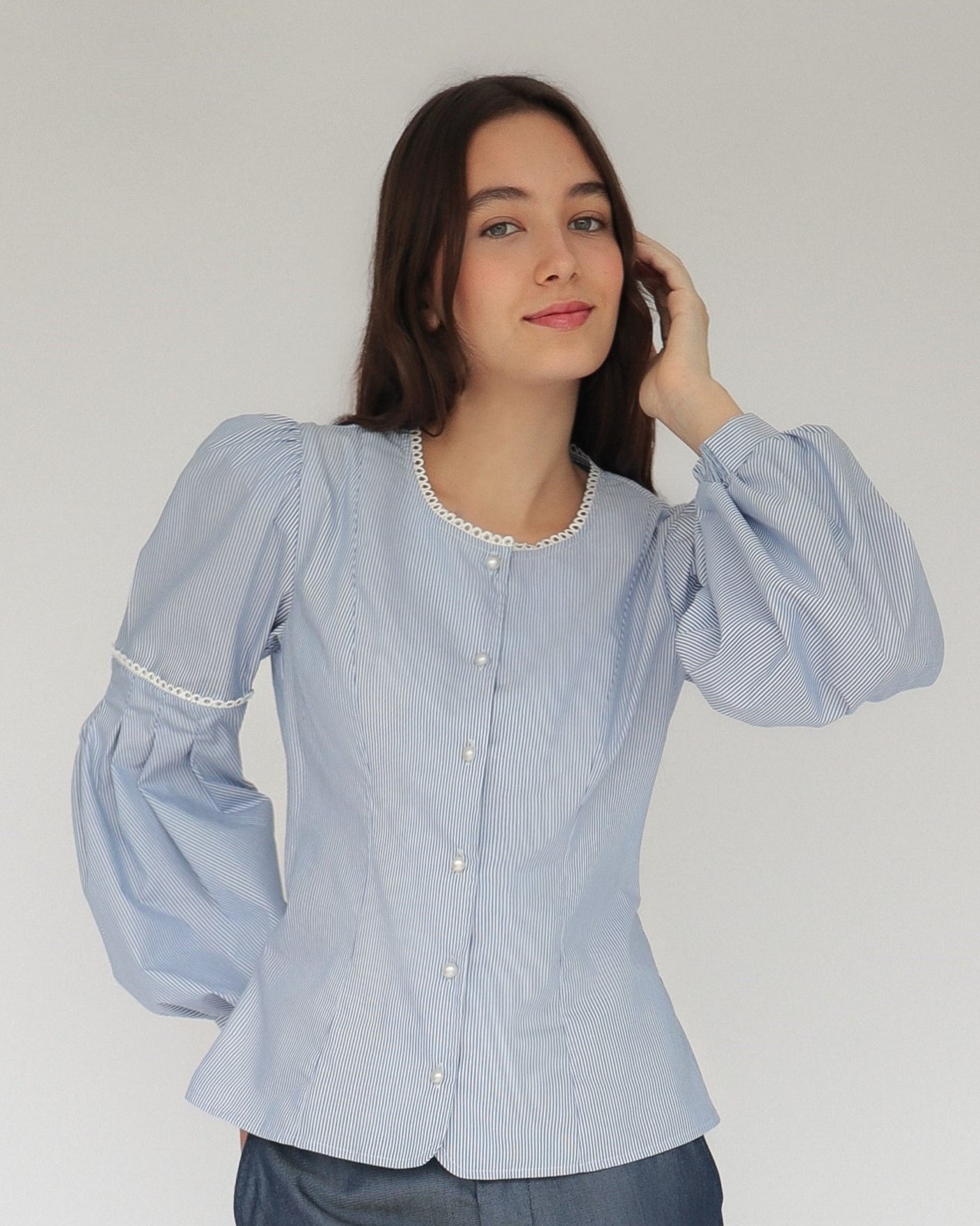Mujer con blusa a rayas celeste con detalles tejidos, cuello redondo y mangas ligeramente voluminosas. Posando en fondo blanco.