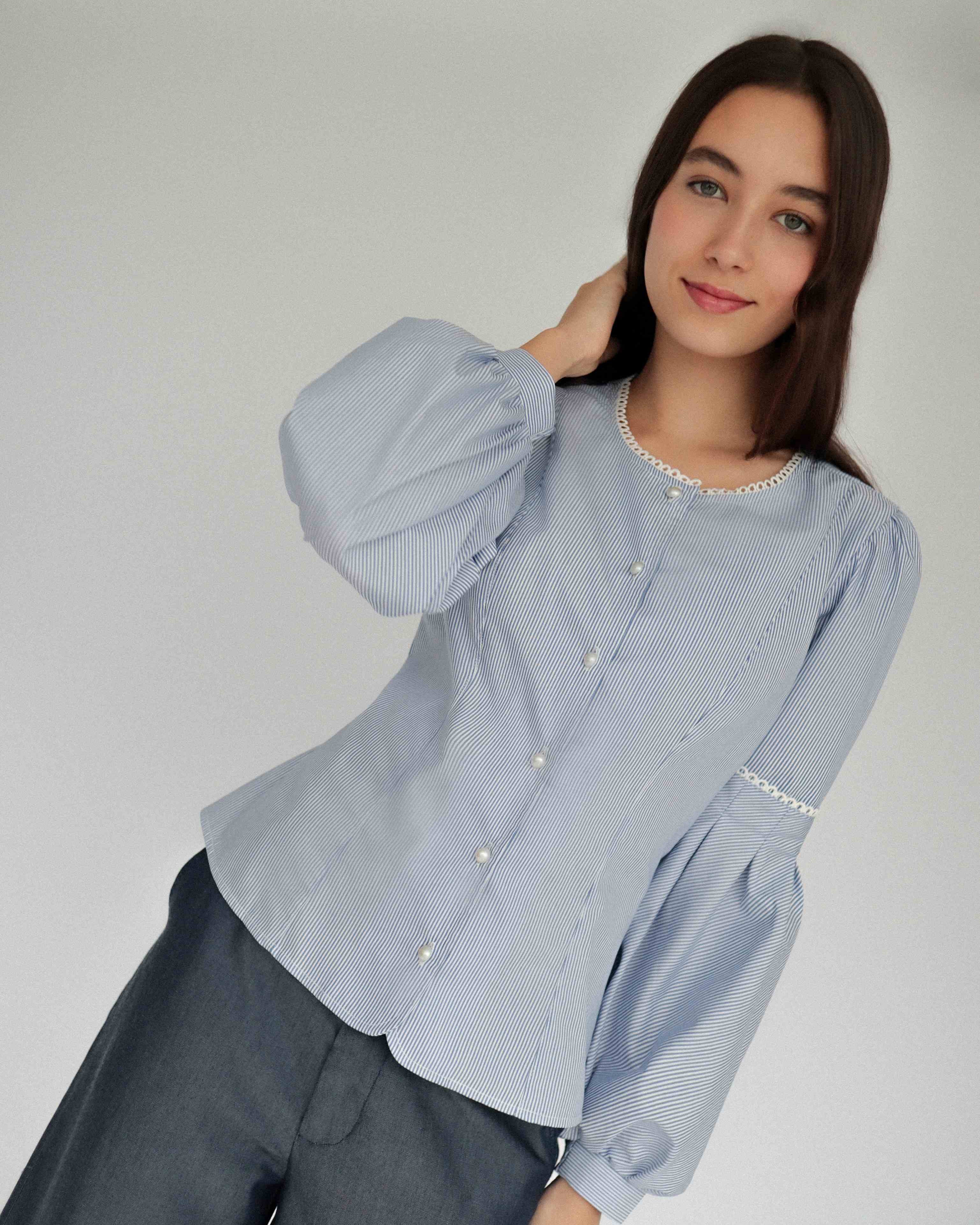 Mujer con blusa a rayas celeste con detalles tejidos, cuello redondo y mangas ligeramente voluminosas. Posando en fondo blanco.