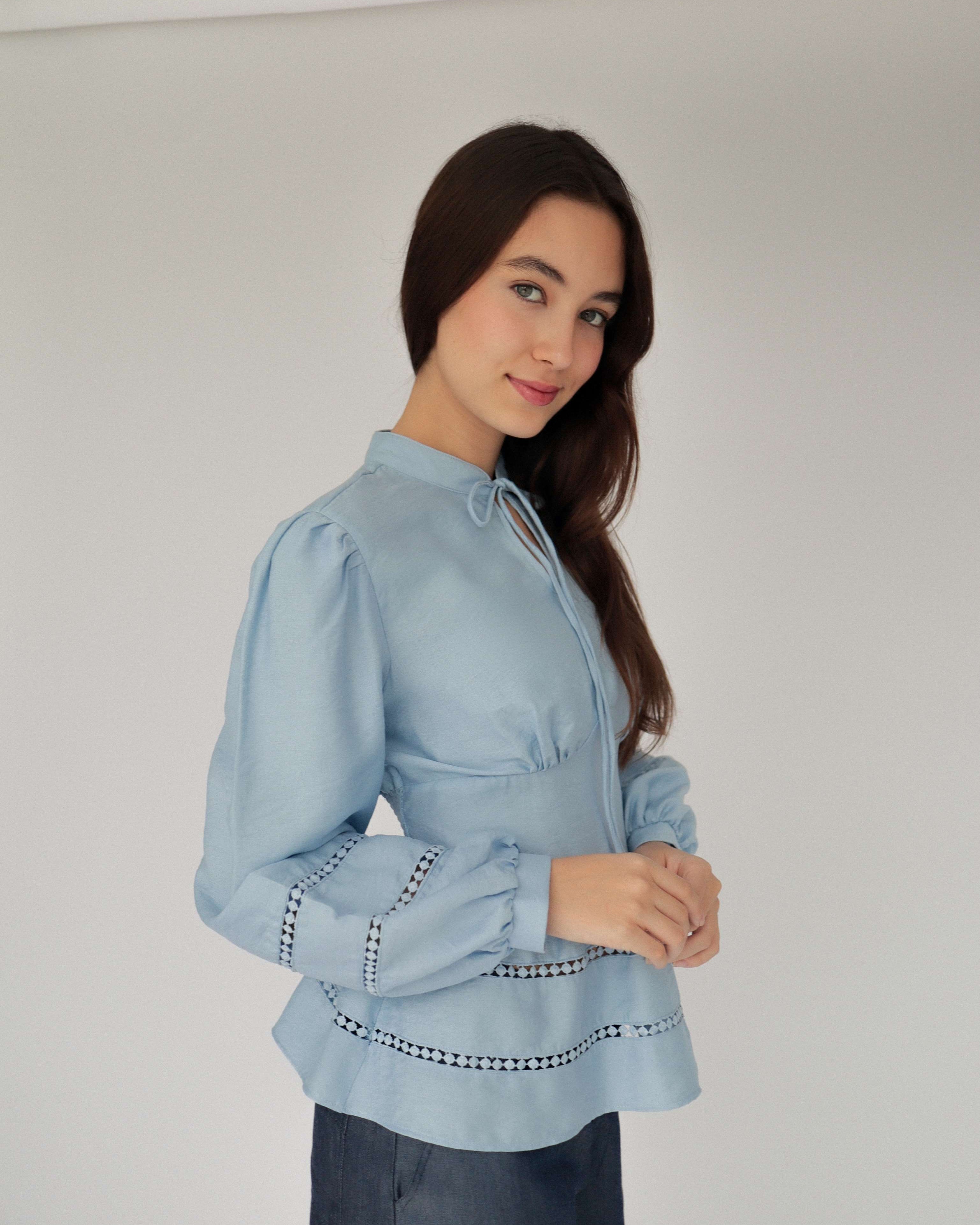 Mujer con blusa celeste con detalles tejidos y pantalones estilo denim posando en fondo blanco.