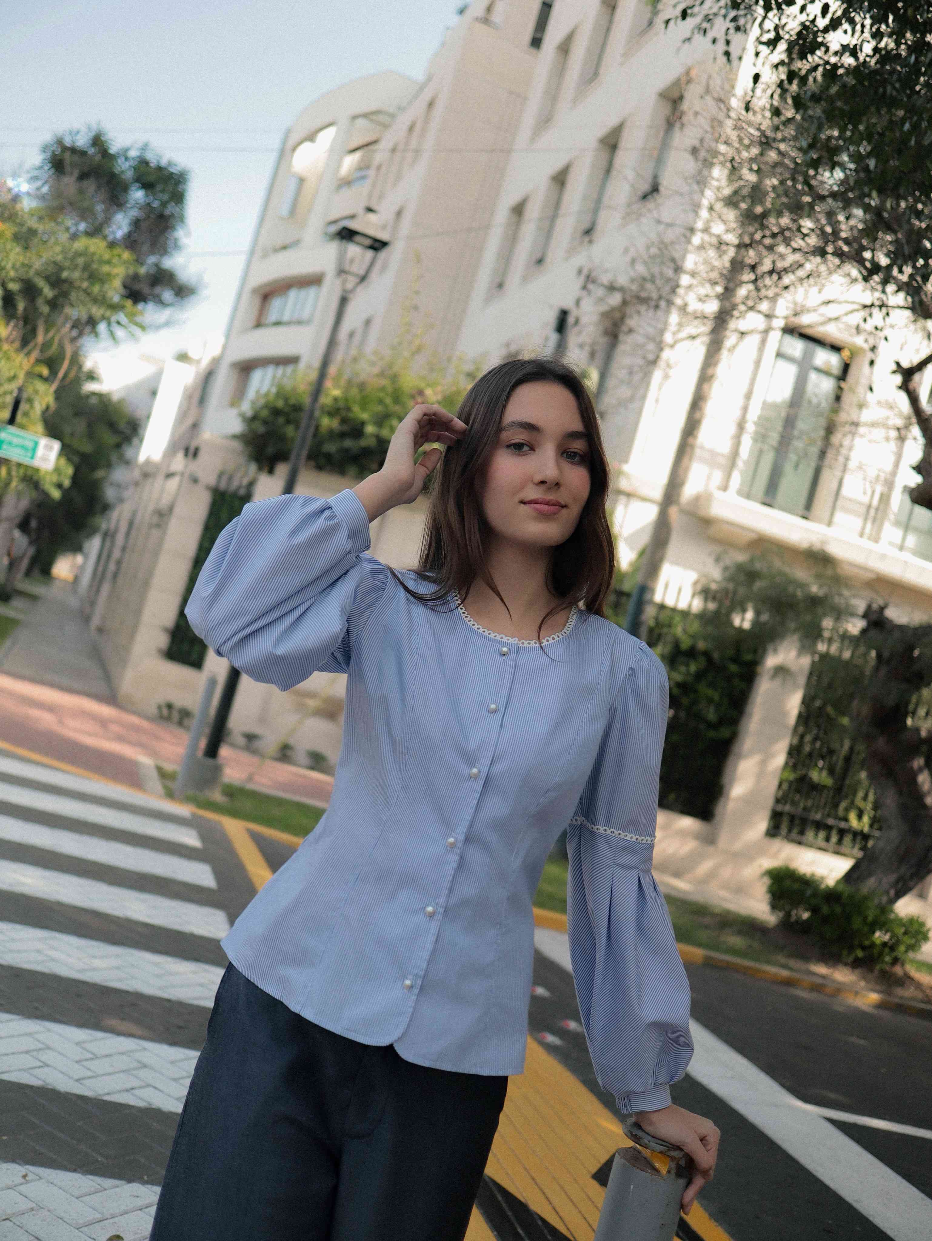 Mujer en la calle con blusa a rayas celeste ceñida al cuerpo con detalles tejidos, cuello redondo y mangas ligeramente voluminosas.