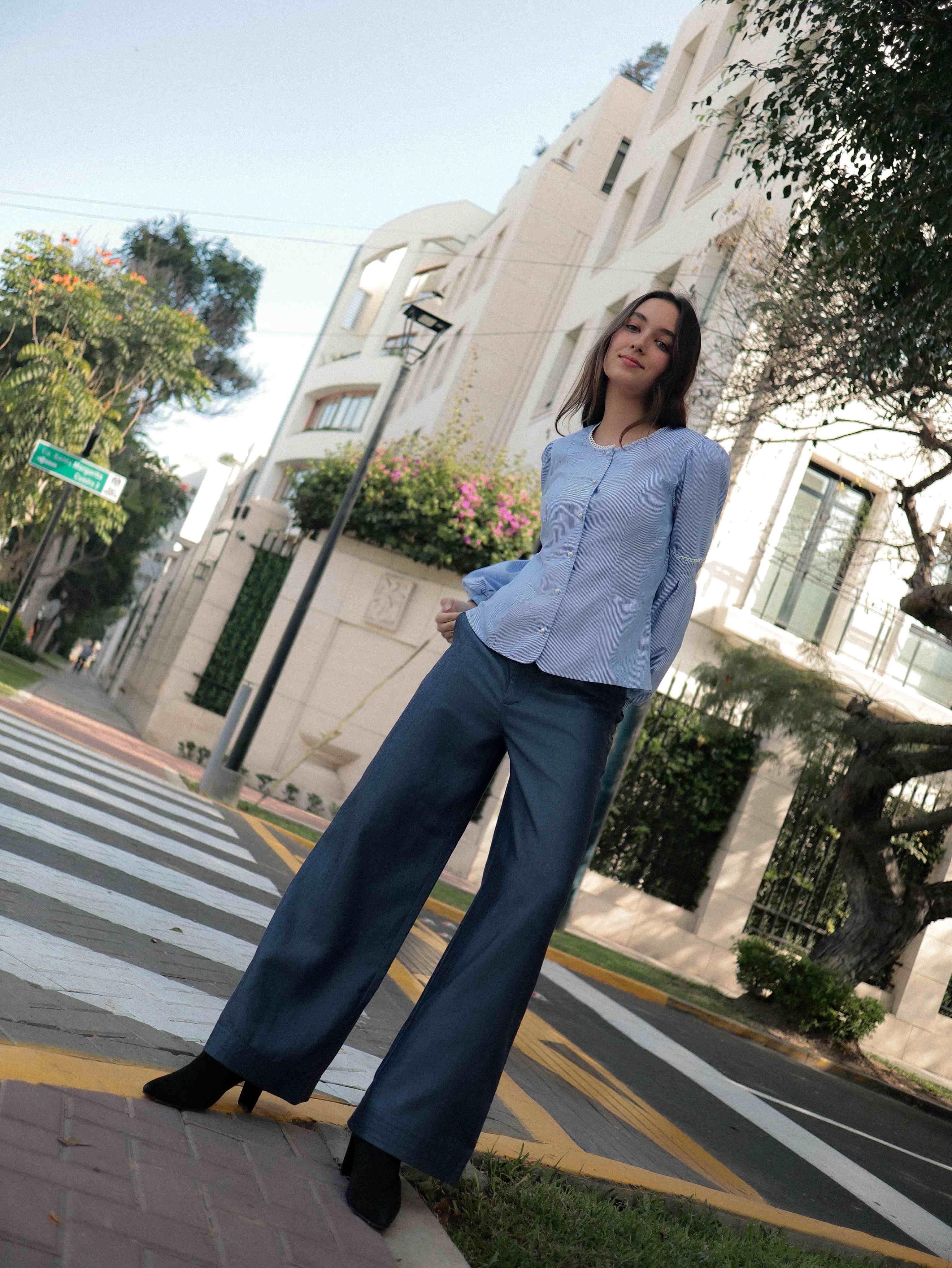Mujer con blusa a rayas celeste con detalles tejidos, cuello redondo y mangas ligeramente voluminosas.  Posando en la calle.
