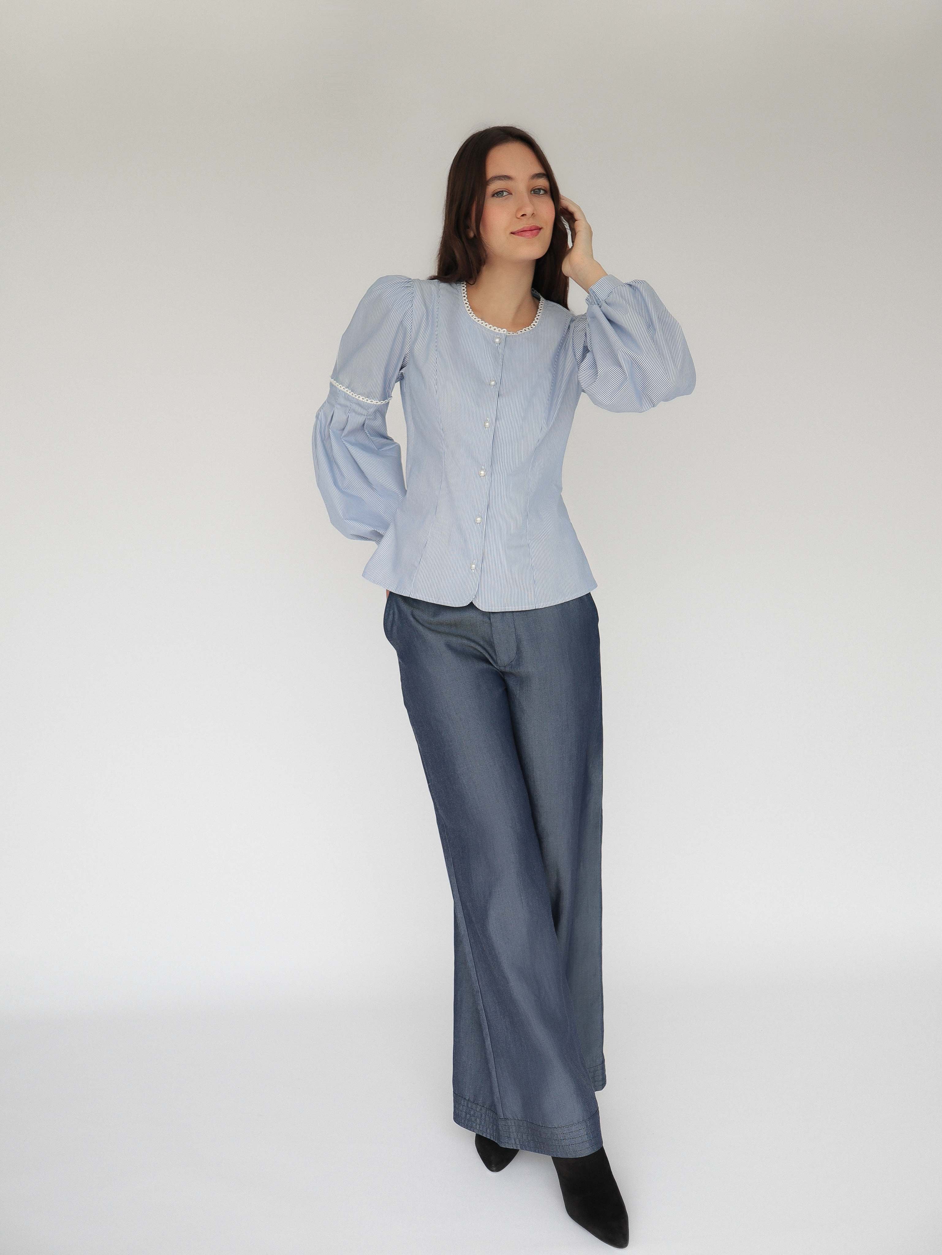 Mujer con blusa a rayas celeste con detalles tejidos, cuello redondo y mangas ligeramente voluminosas.  Posando en fondo blanco.
