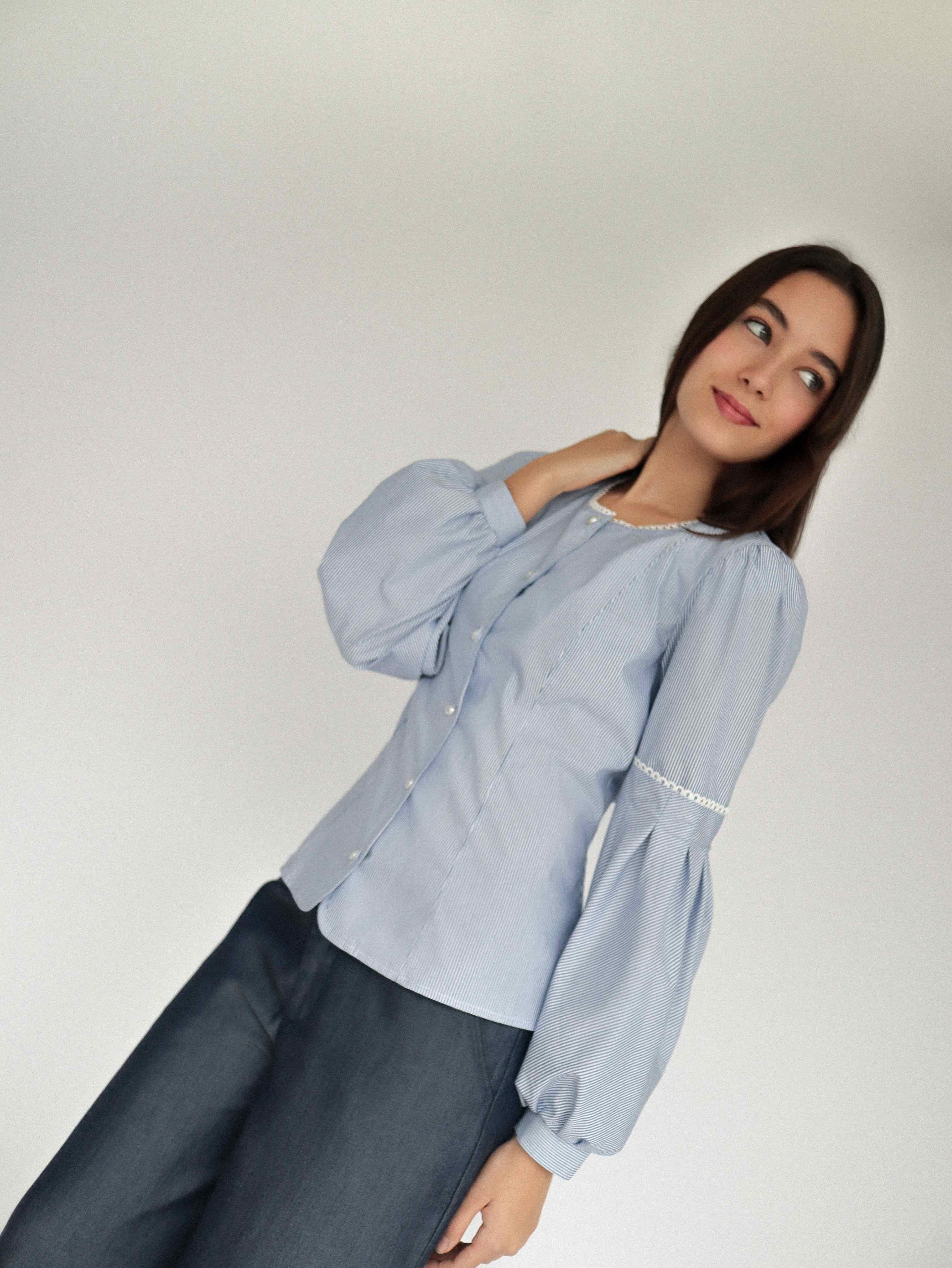 Mujer con blusa a rayas celeste con detalles tejidos, cuello redondo y mangas ligeramente voluminosas.  Posando en fondo blanco.