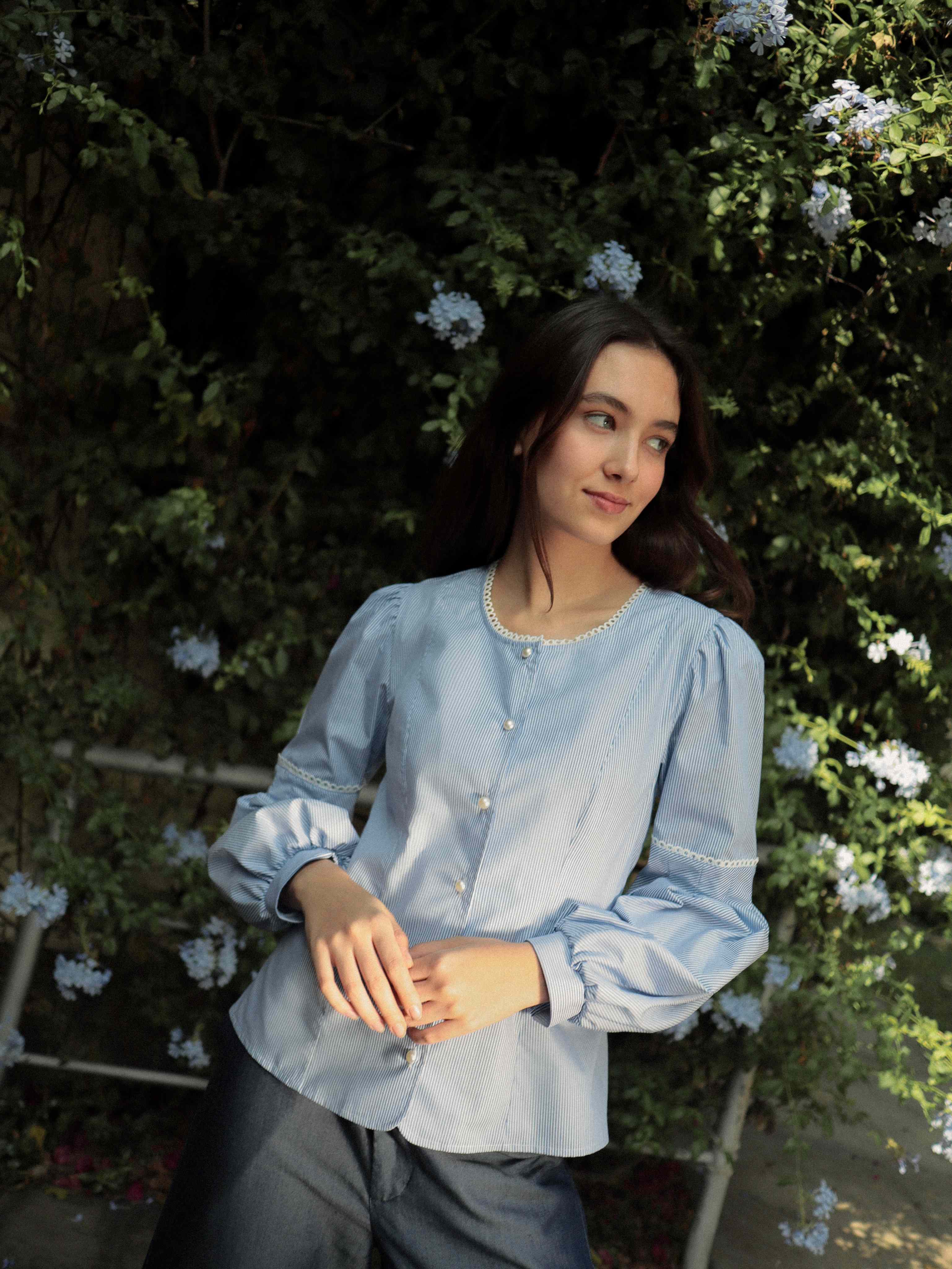 Mujer con blusa a rayas celeste con detalles tejidos, cuello redondo y mangas ligeramente voluminosas.  Posando en fondo de flores.