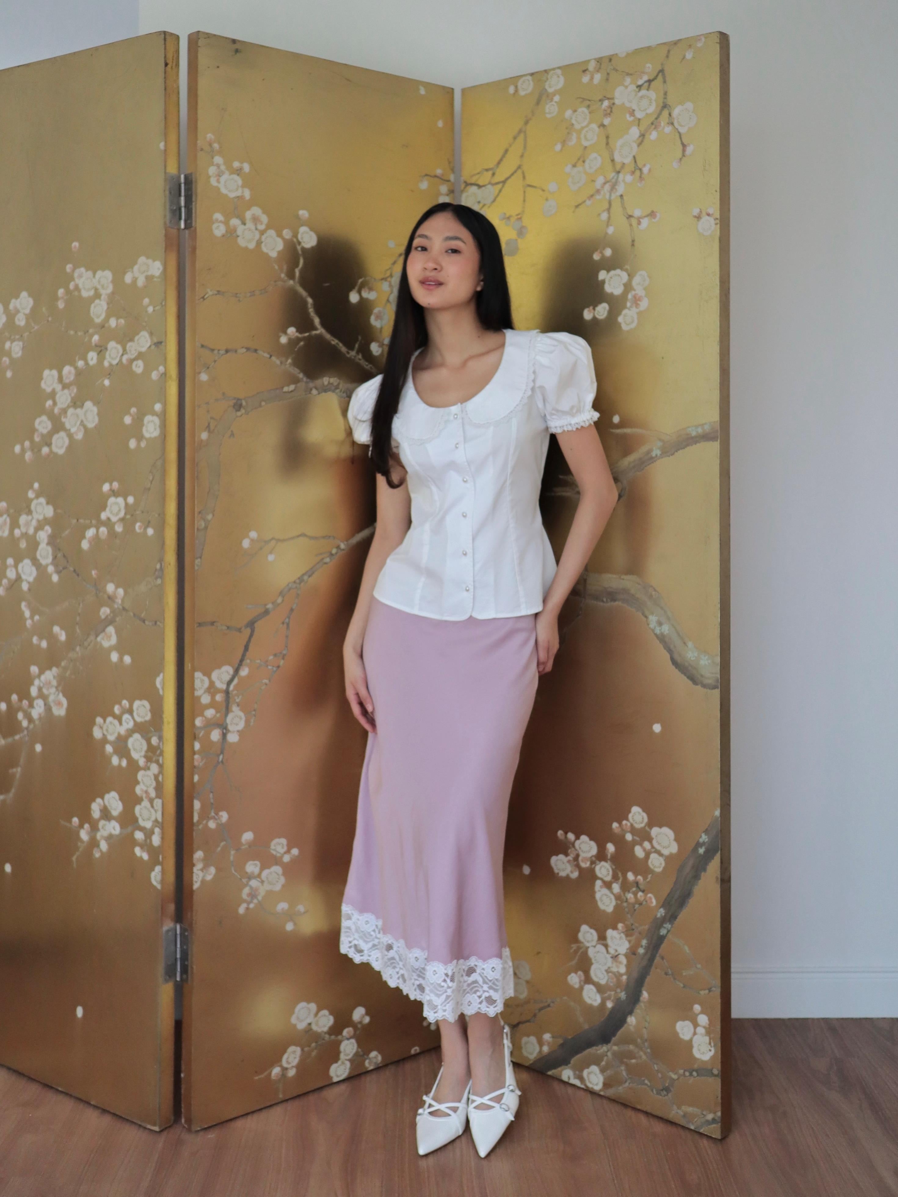 Mujer con blusa blanca de algodón romántica y falda rosa midi. Posando en fondo dorado con diseño floral