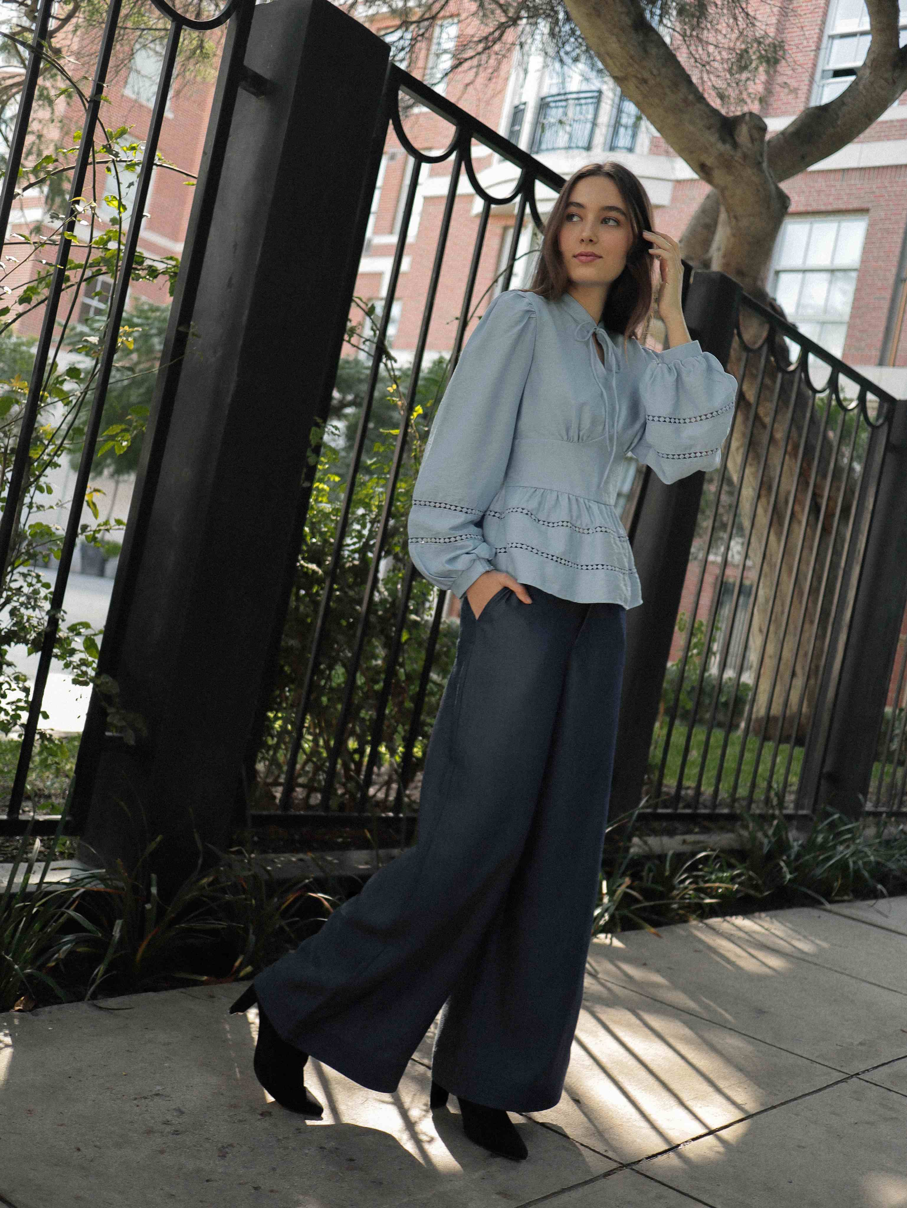 Mujer joven con blusa celeste con detalles tejidos, pantalones estilo denim y botas negras posando en fondo blanco