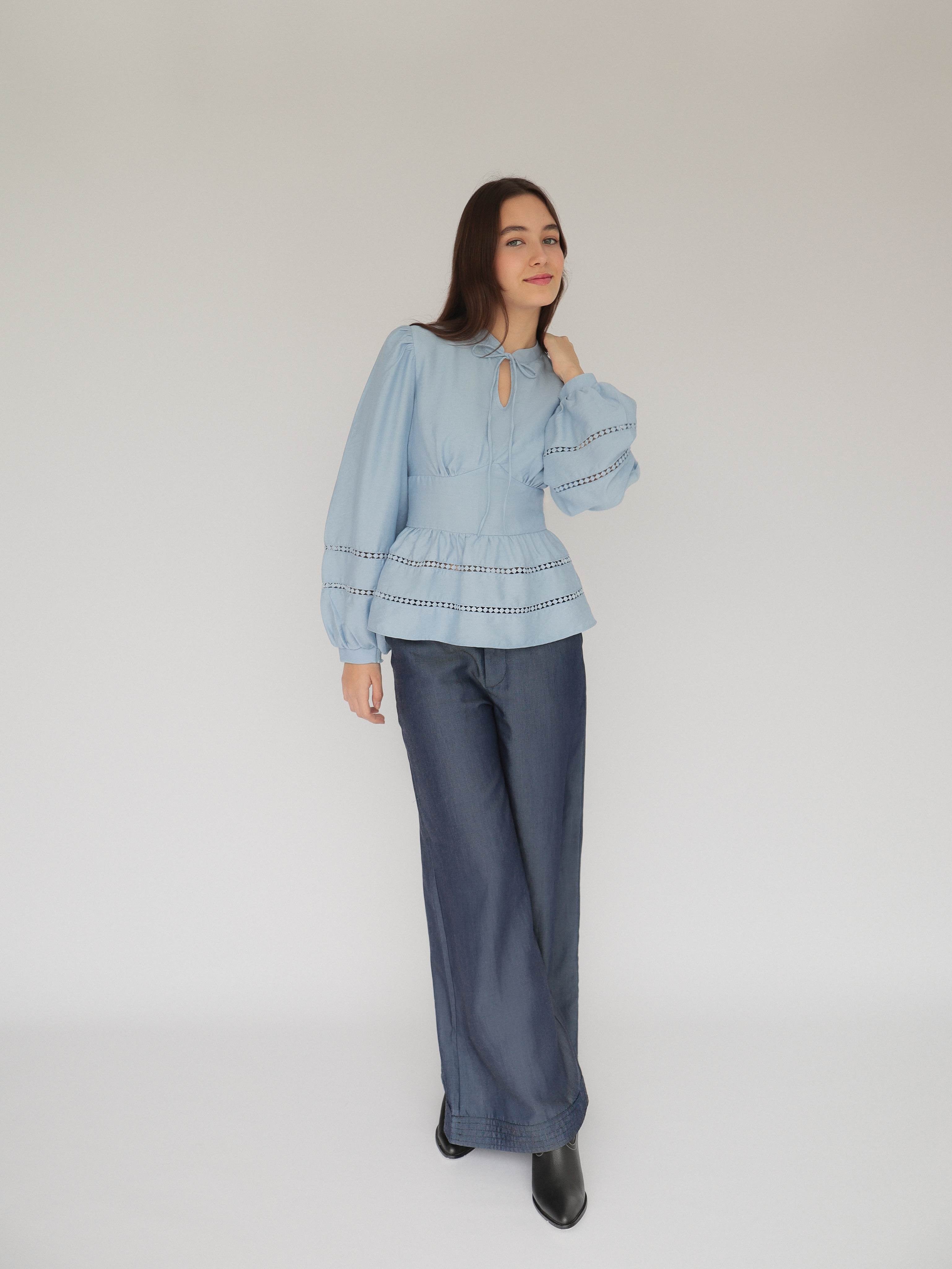 Mujer joven con blusa celeste con detalles tejidos, pantalones estilo denim y botas negras posando en fondo blanco