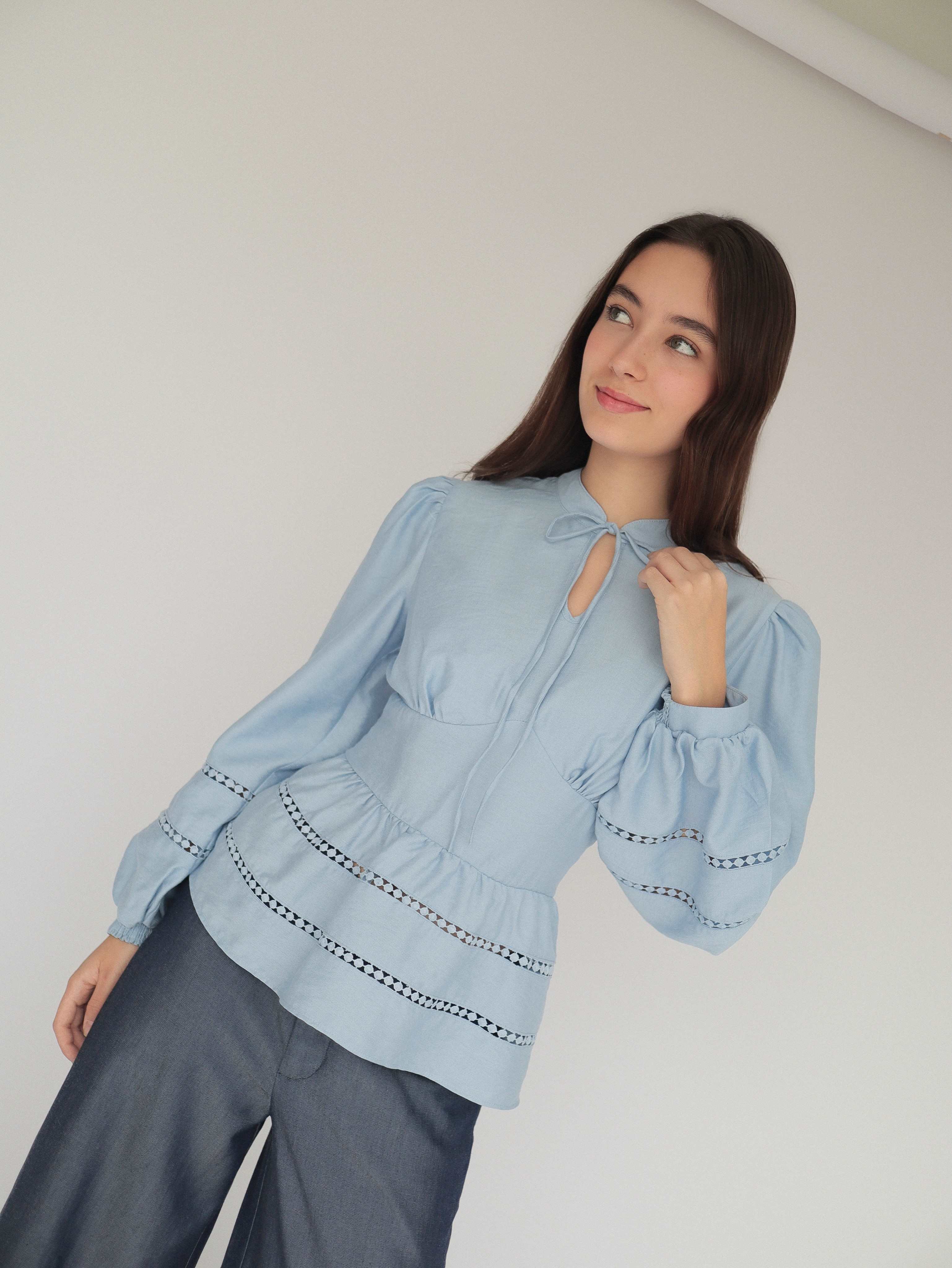 Mujer joven con blusa celeste con detalles tejidos y pantalones estilo denim posando en fondo blanco