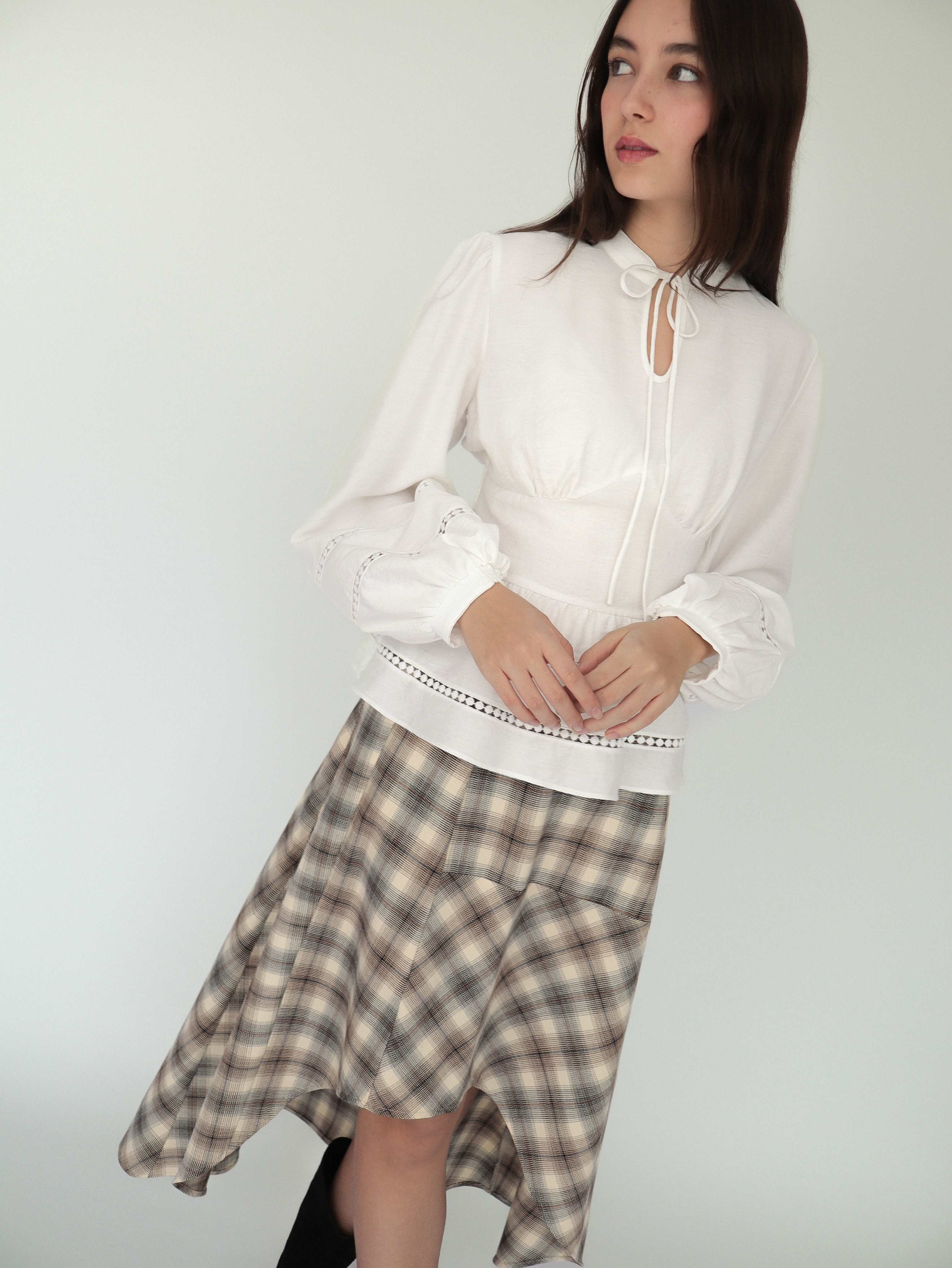 Mujer con blusa blanca sofisticada y elegante estilo peplum ceñida, con cuello alto y detalles tejidos. Lo combina con falda asimétrica a cuadros.