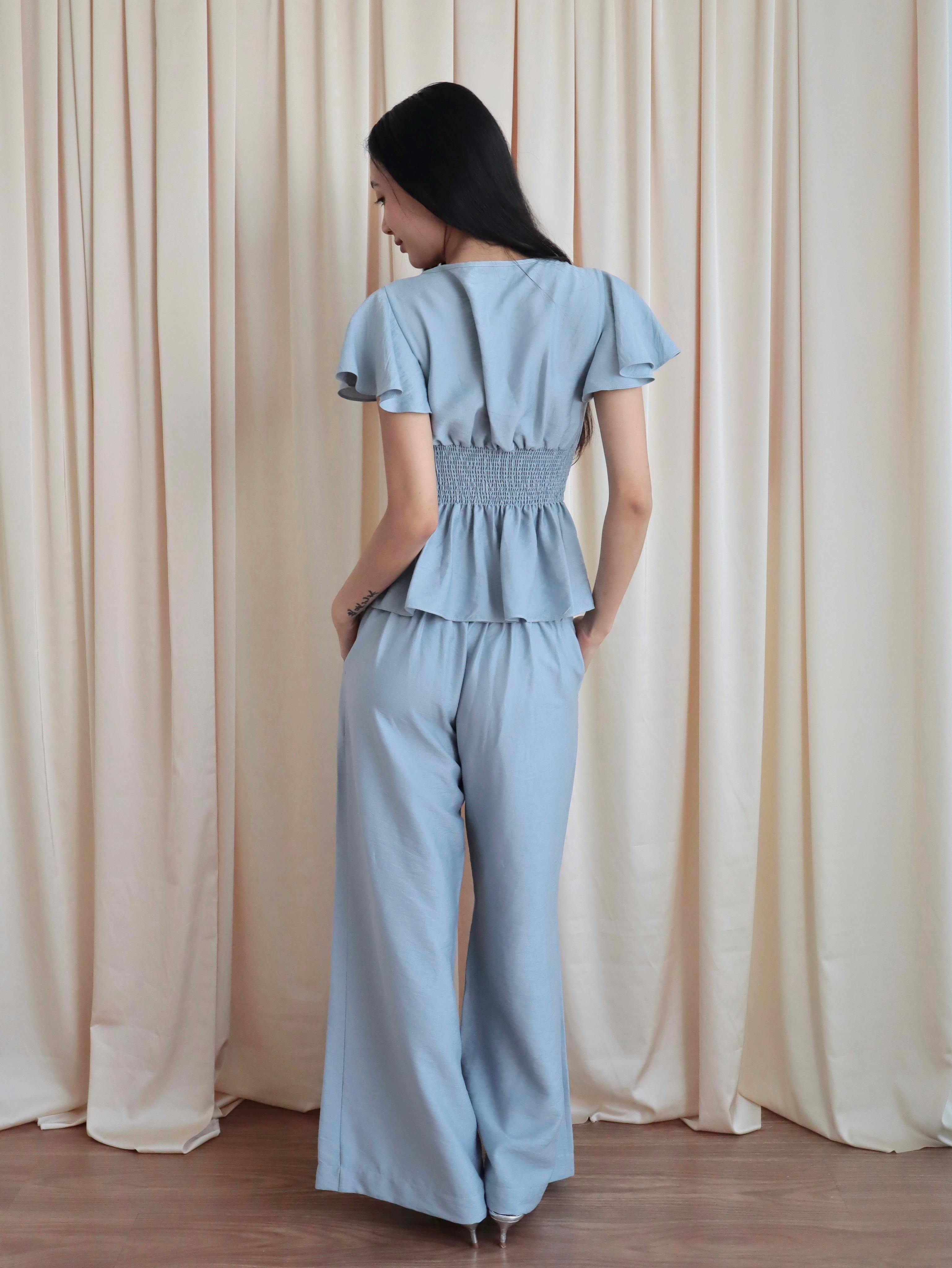 Mujer de espaldas usando blusa celeste romántica con bobos en mangas, lazo y corte que marca la cintura. Usa un pantalón recto celeste. Posando en fondo amarillo pastel.