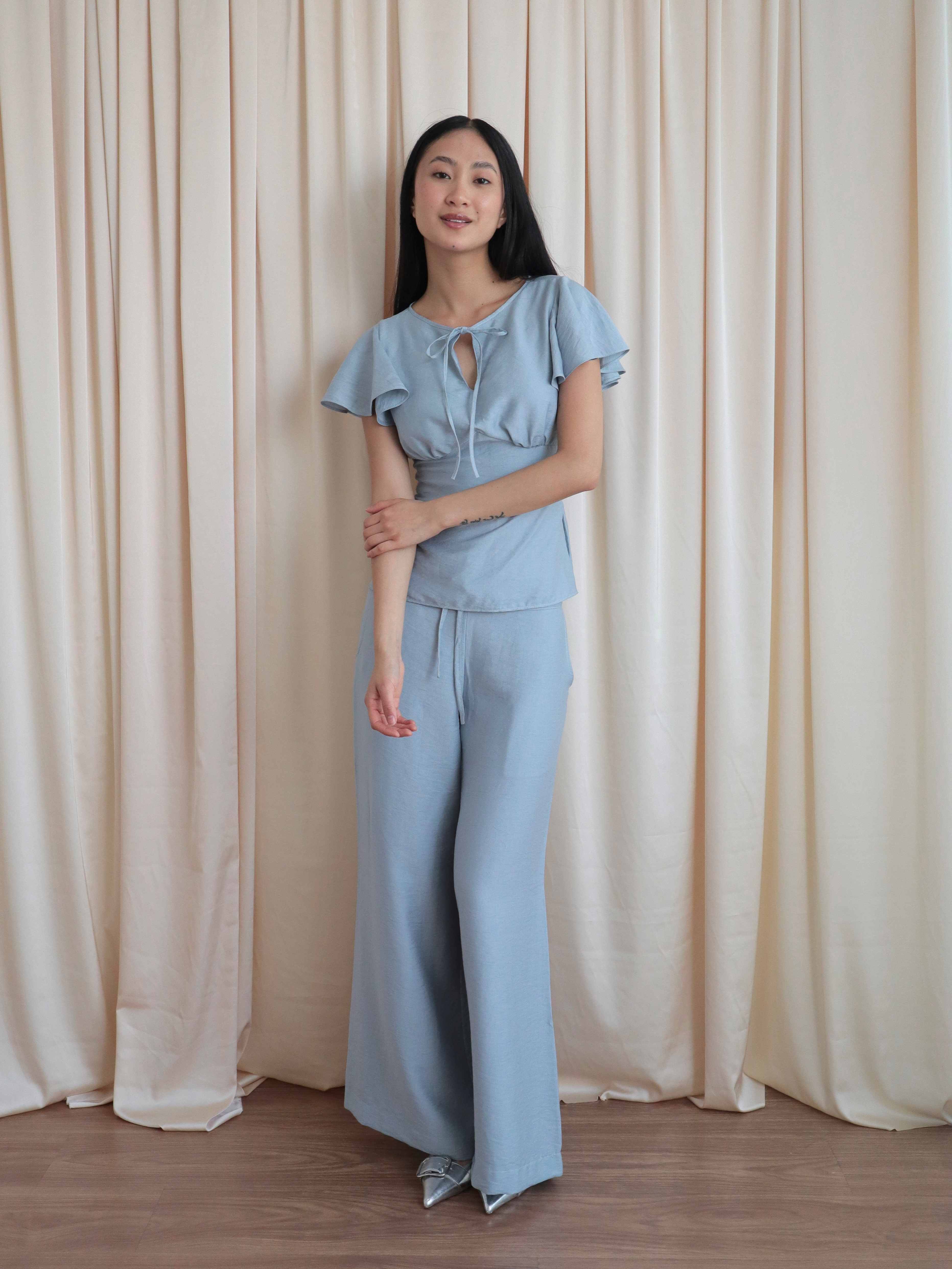 Mujer con blusa celeste romántica con bobos en mangas y lazo. Pantalón recto celeste. Posando en fondo amarillo pastel.