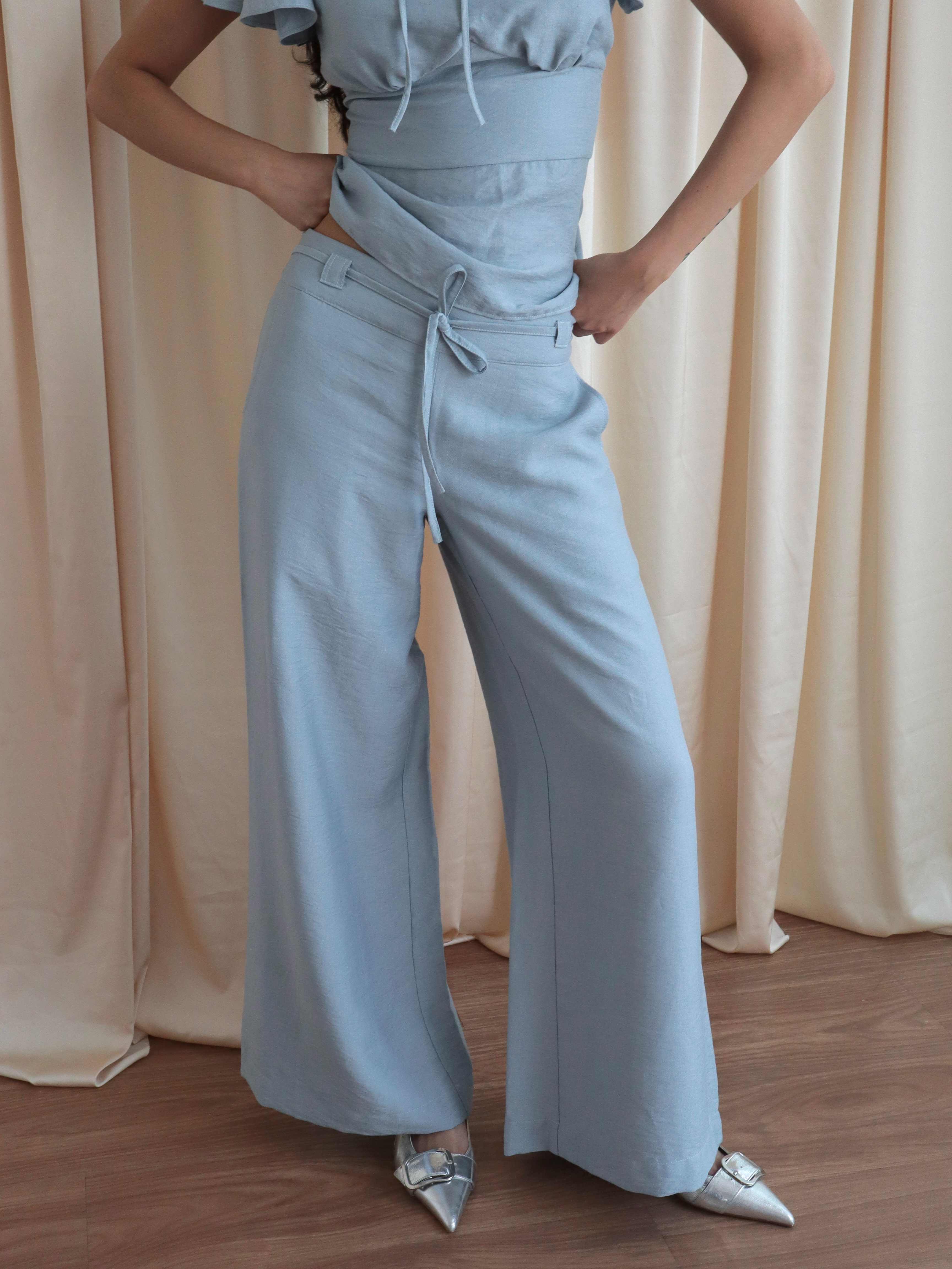 Mujer usando pantalón recto a la cadera celeste con lazo en pretina. Posando en fondo amarillo pastel.