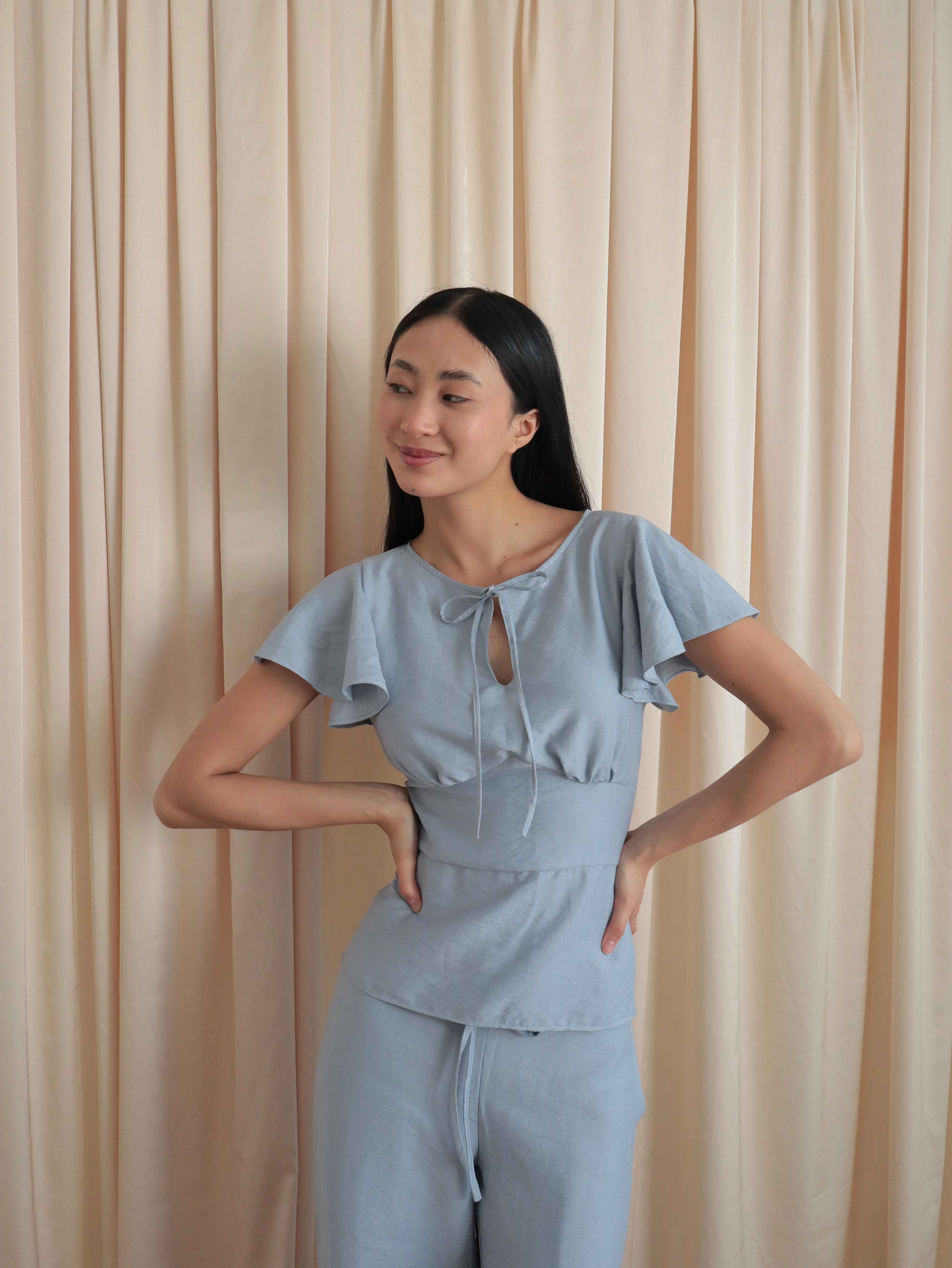 Mujer con blusa celeste romántica con bobos en mangas, lazo y corte que marca la cintura. Posando en fondo amarillo pastel.