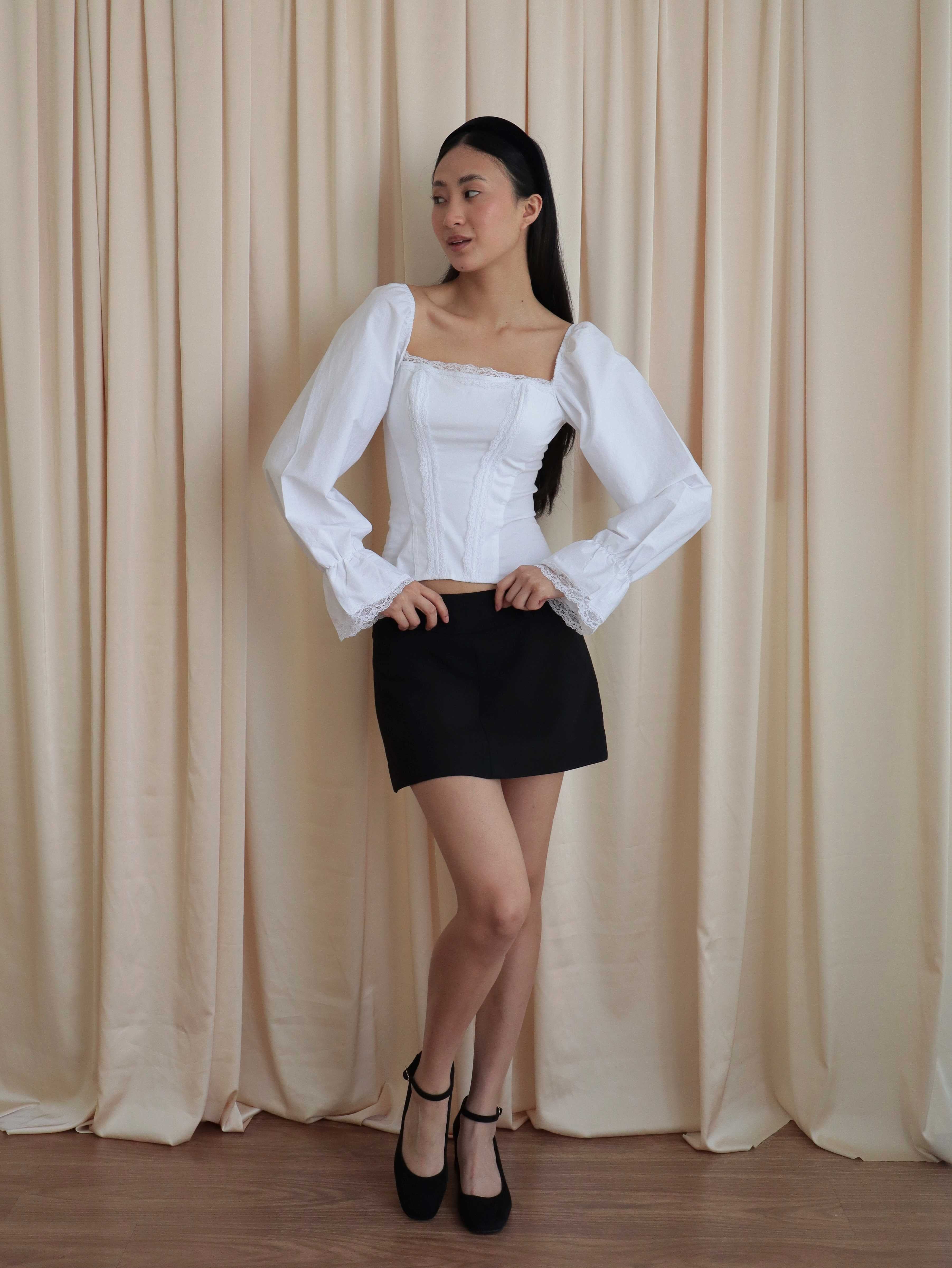 Mujer usando corset de algodón, mangas largas y blondas, usando falda negra corta con ballerinas. Posando en fondo amarillo pastel.