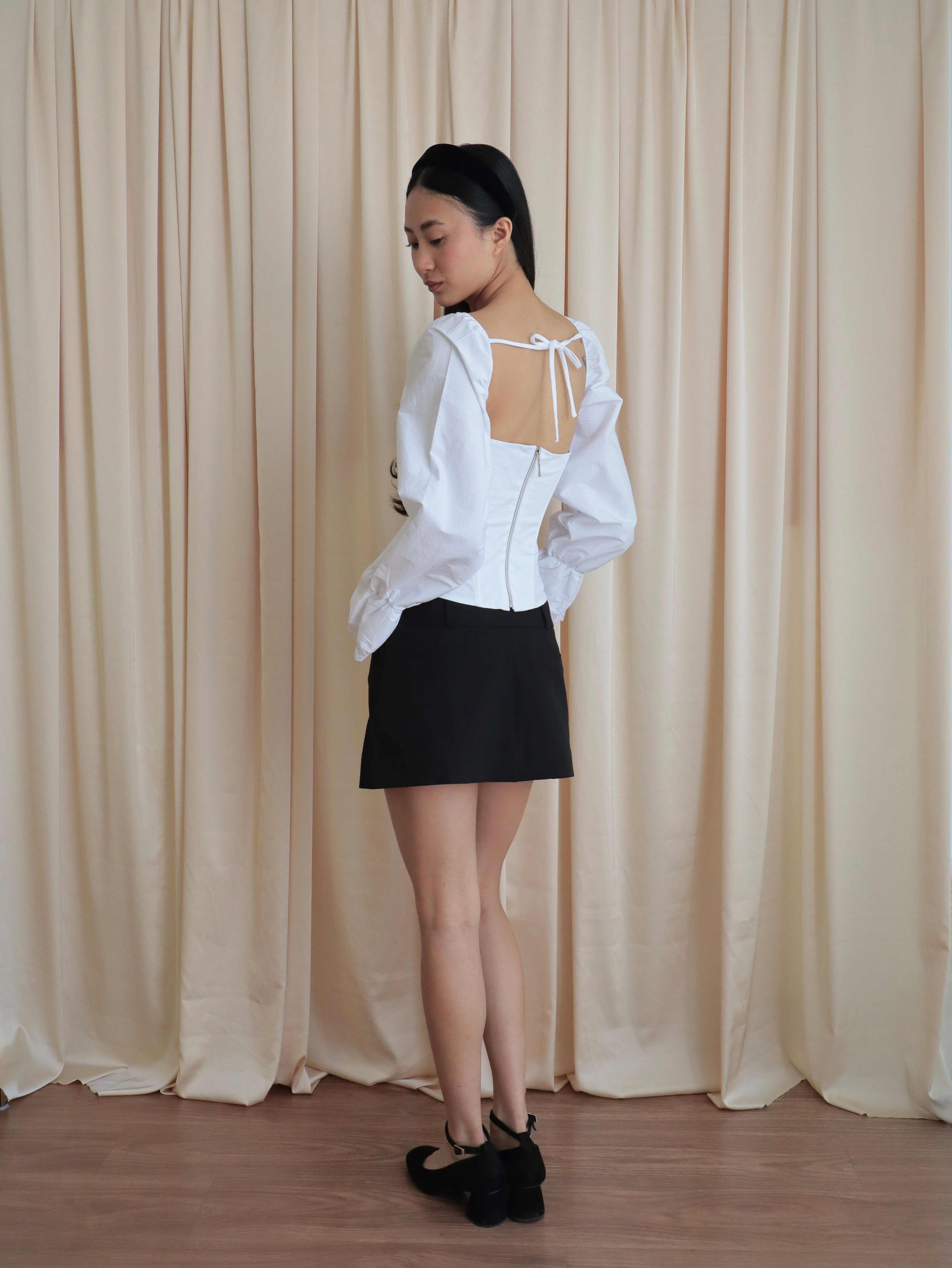 Mujer usando corset de algodón, mangas largas y blondas, usando falda negra corta con ballerinas. Posando en fondo amarillo pastel.