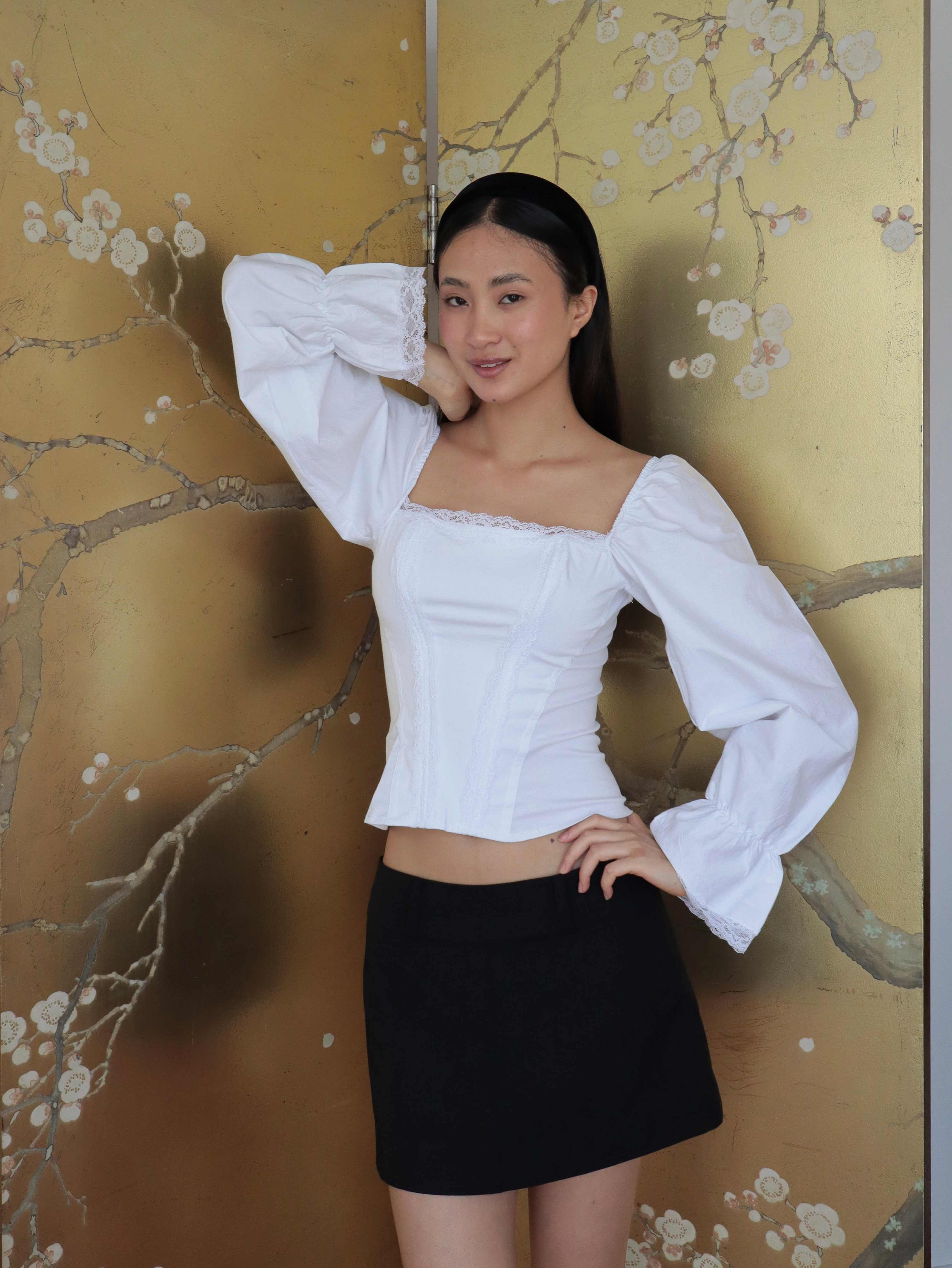 Mujer usando corset de algodón, mangas largas y blondas, usando falda negra corta. Posando en fondo floral.