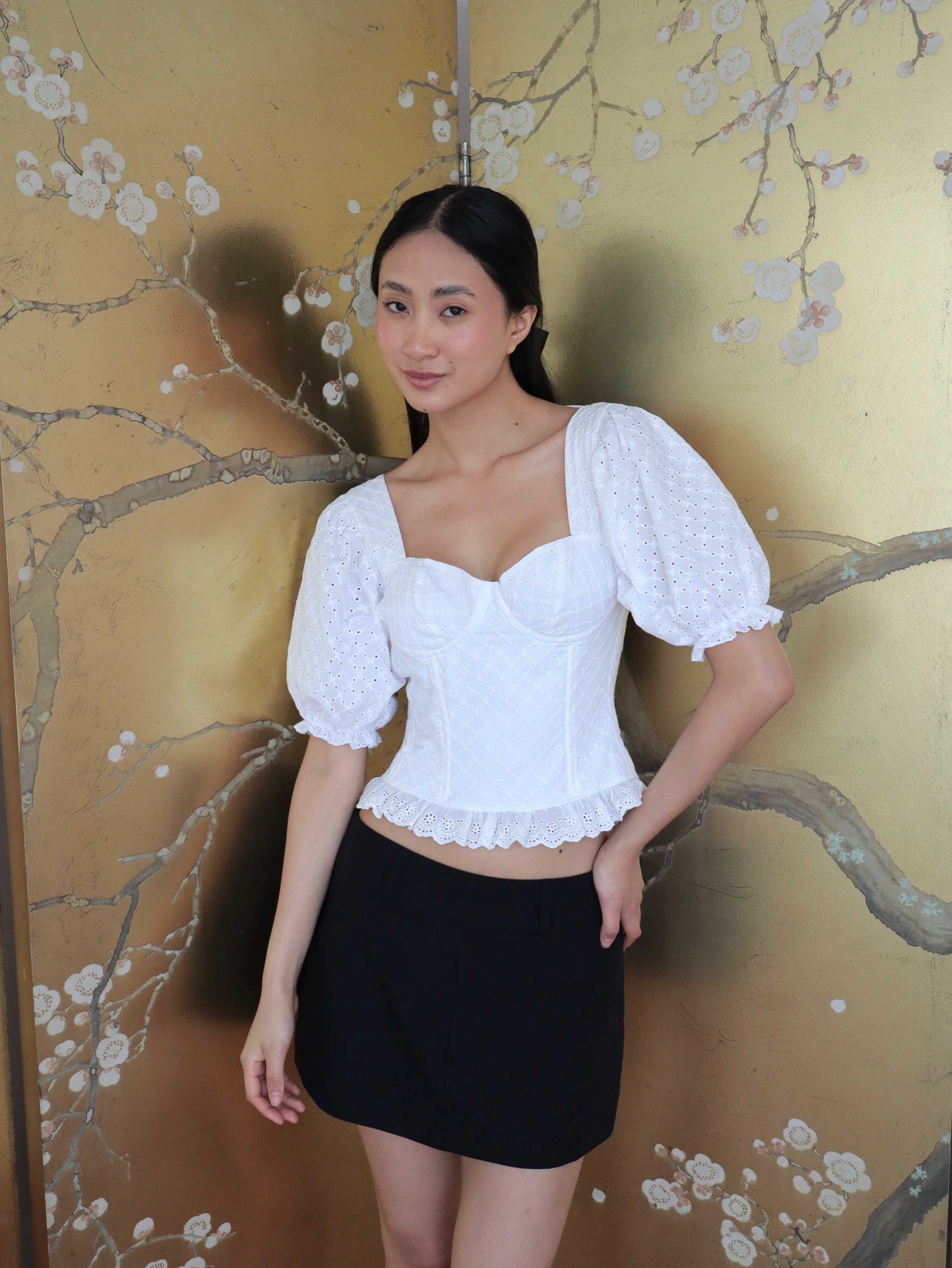 Mujer con corset broderie con escote corazón, mangas abullonadas y blonda usando falda negra corta. Posando en fondo floral.